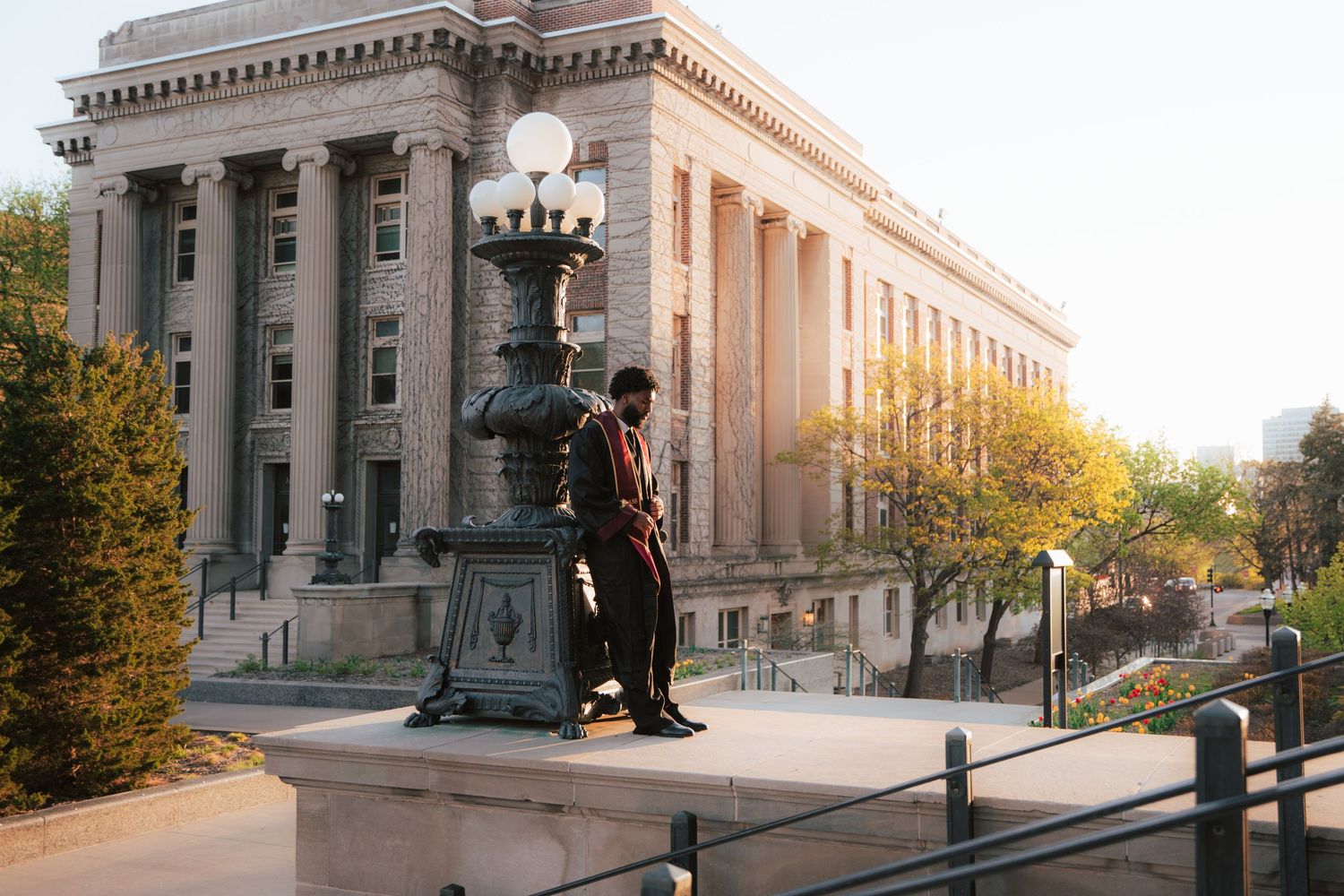 Man in formal attire leans against ornate lamppost outside a grand neoclassical building at golden hour.