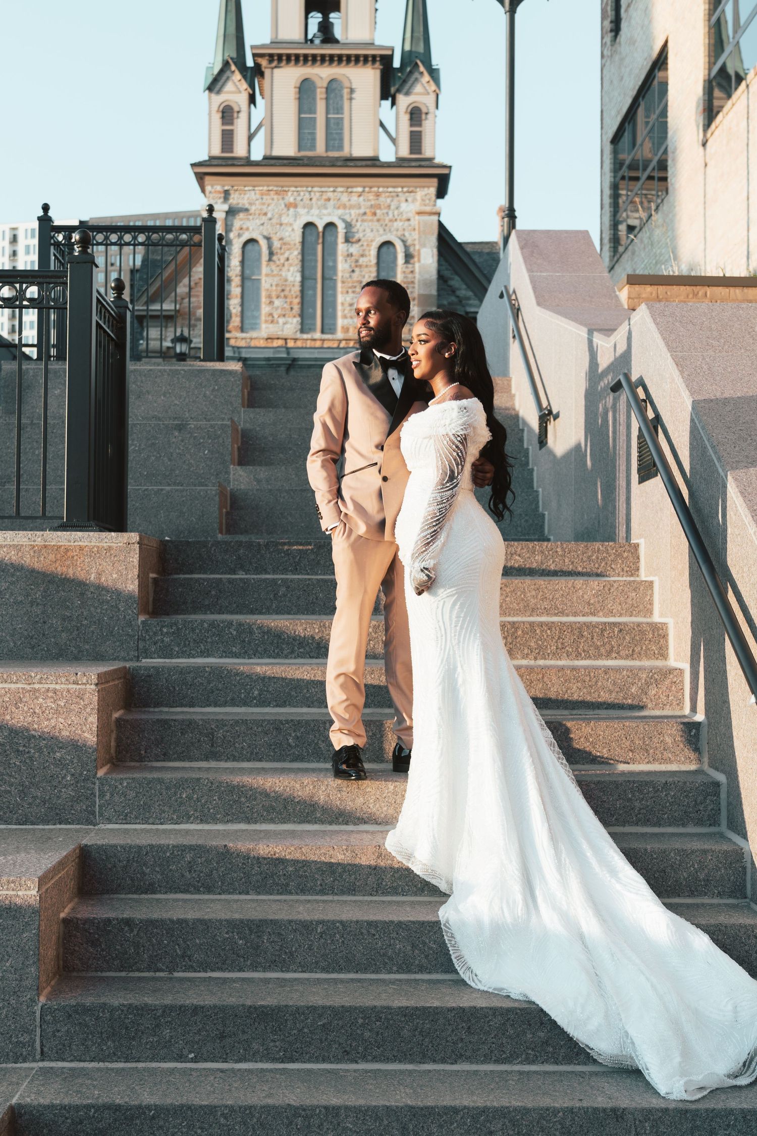 Elegant couple posing on stone steps, groom in beige suit and bride in white off-shoulder mermaid wedding gown.