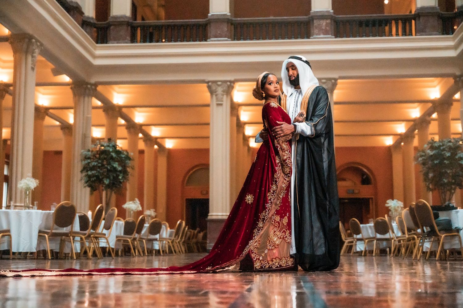 Couple in traditional Middle Eastern wedding attire posing in an elegant ballroom with marble floors.