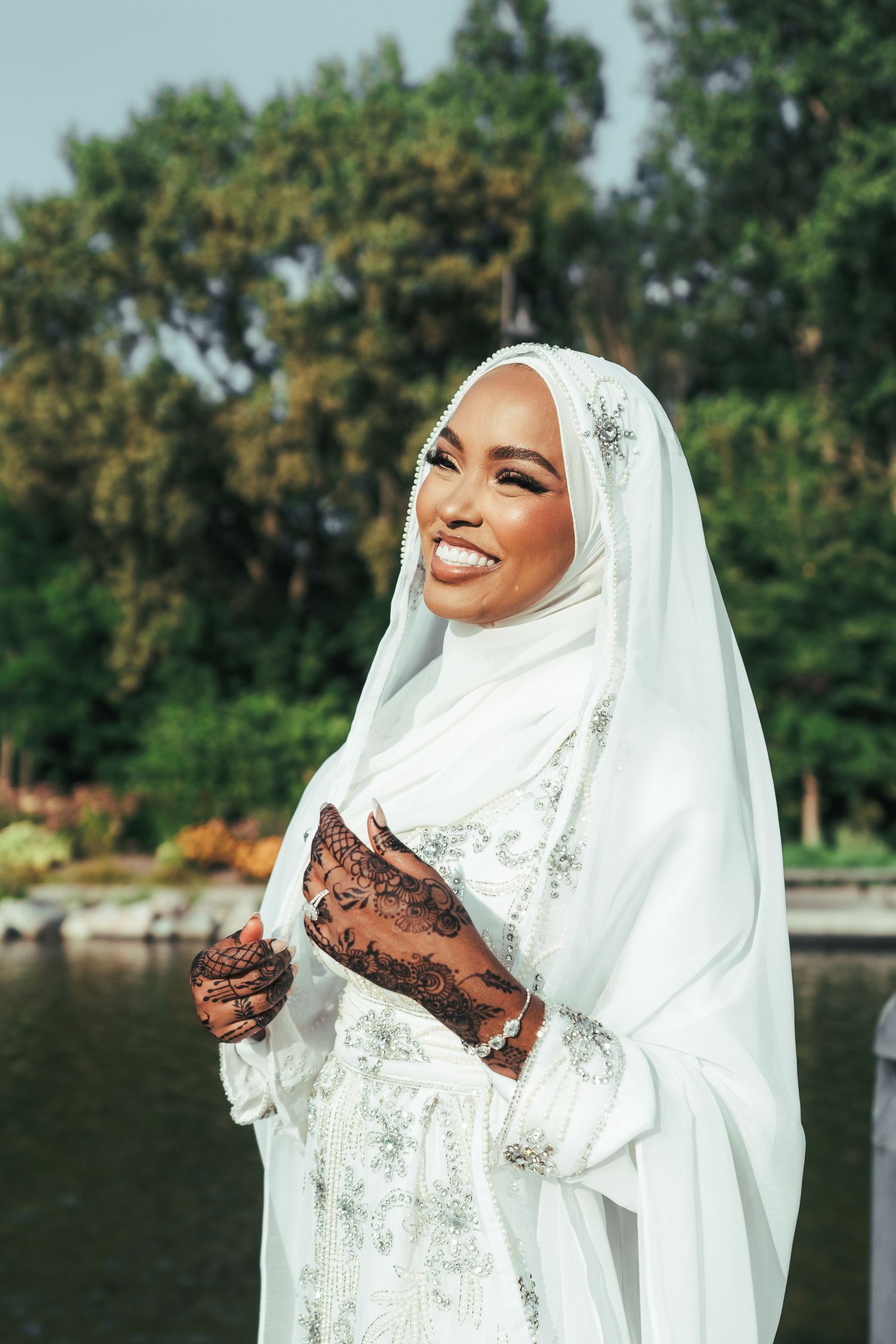 Smiling Muslim bride in white hijab bridal gown with henna hands poses near water in park.