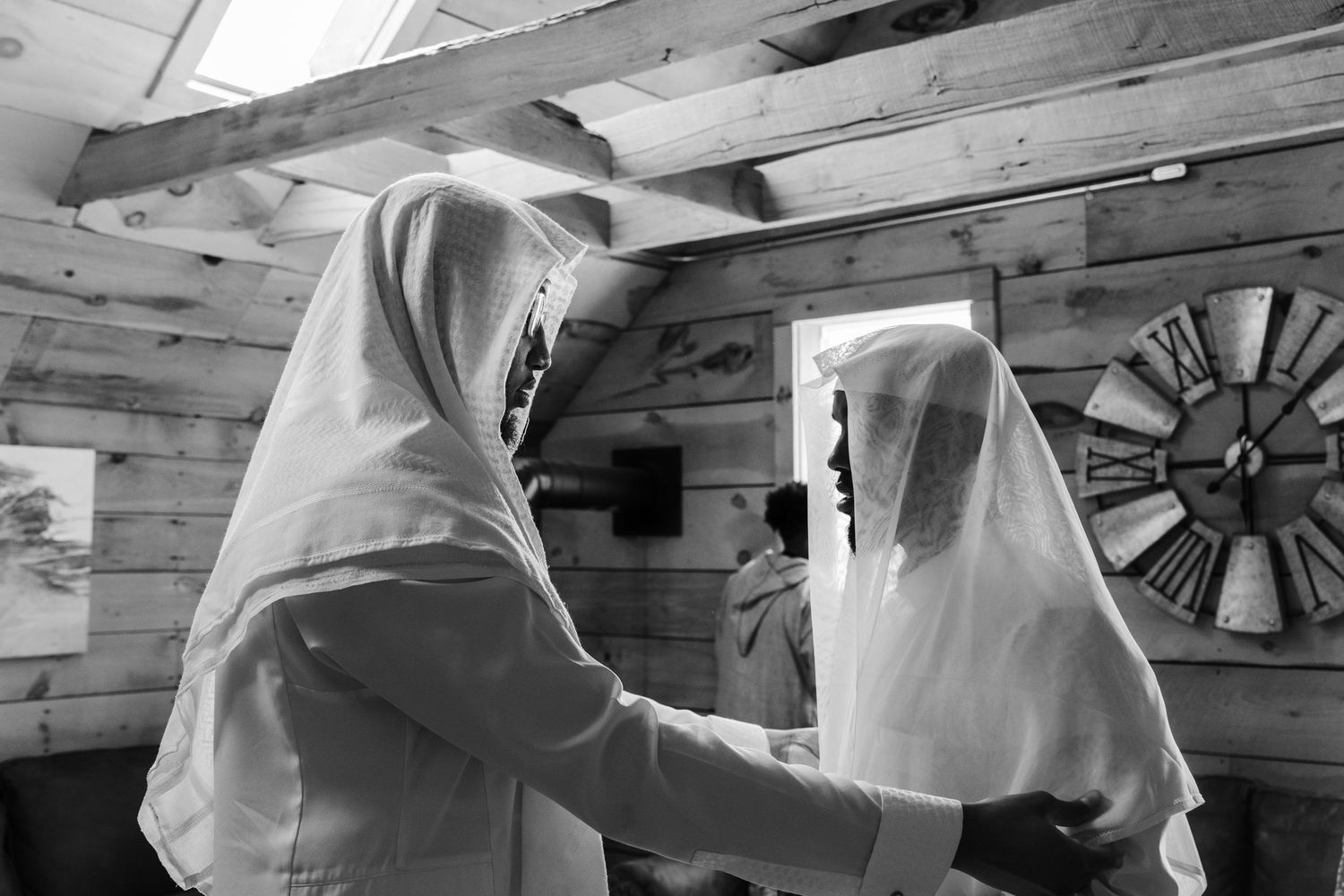 Two women in white hijabs help each other prepare in a rustic wooden cabin during a Muslim wedding ceremony.