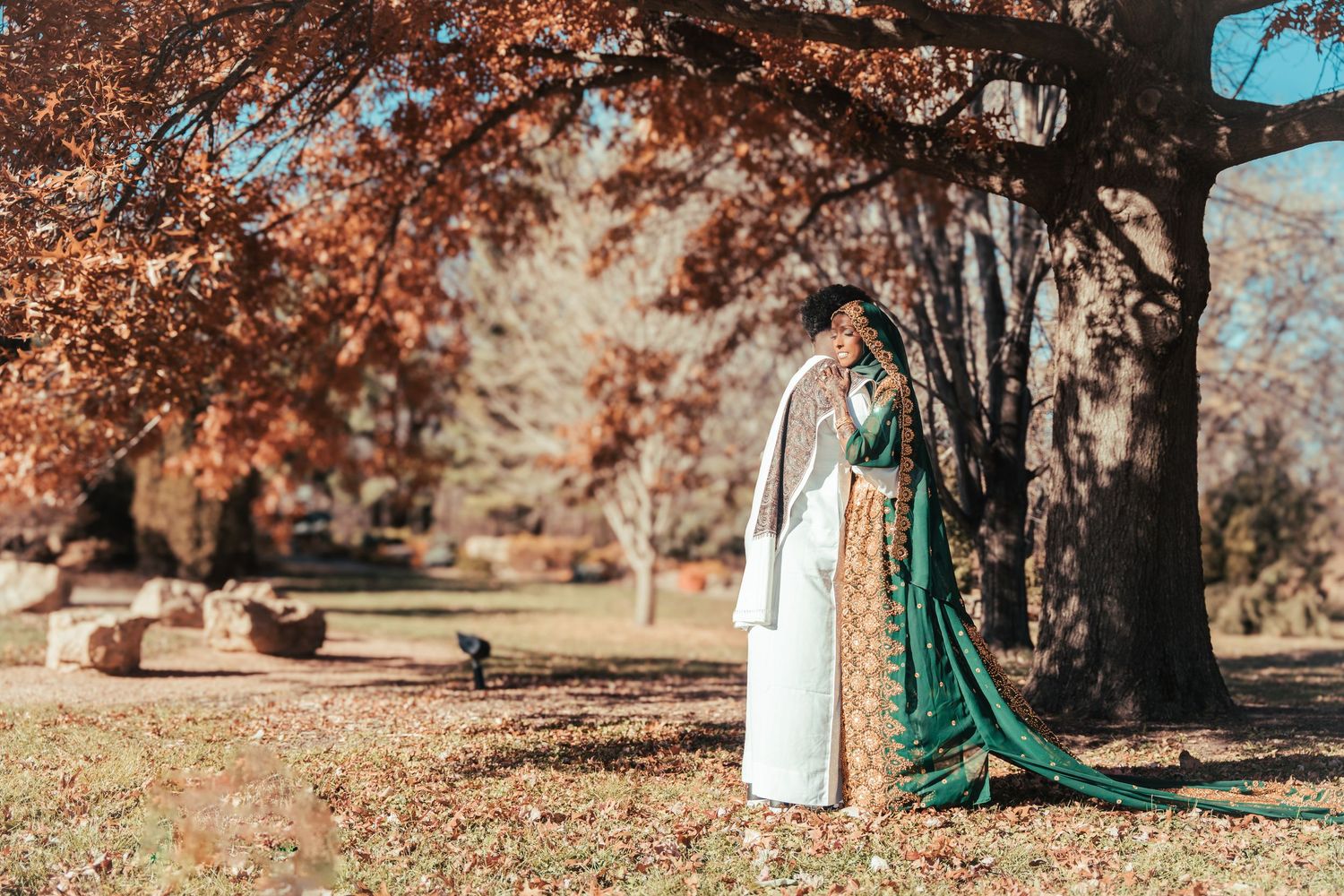 Bride in green and white traditional bridal outfit standing under autumn trees in a park.