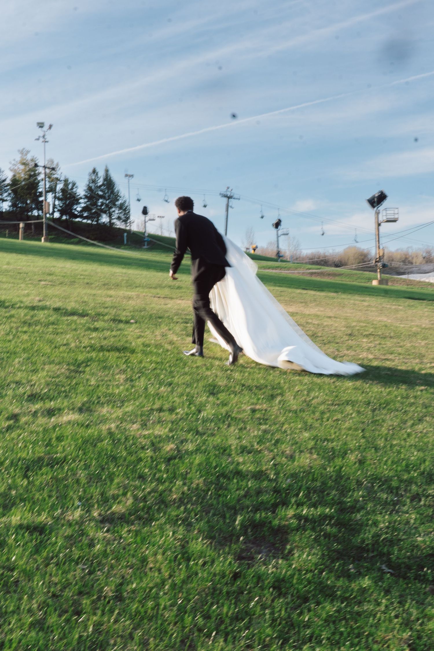 A groom in black carries a bride's flowing white wedding dress train across a green outdoor field.