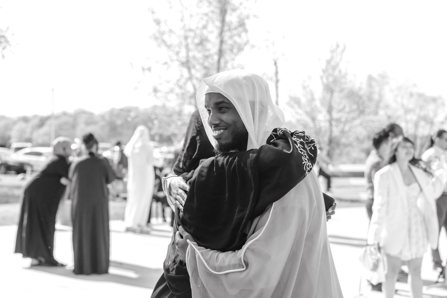 Two people sharing a warm embrace outdoors at a gathering in traditional Islamic clothing.