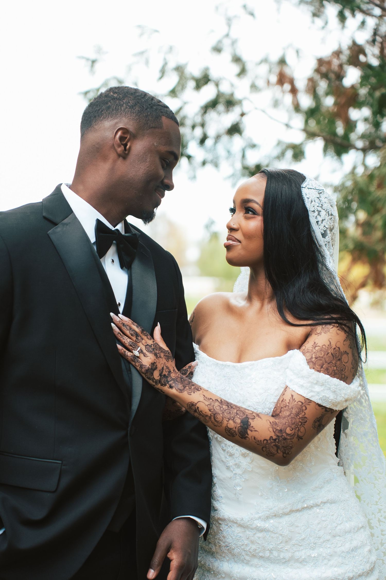 A Black bride in an off-shoulder gown and veil gazes at her groom in a tuxedo outdoors on their wedding day.