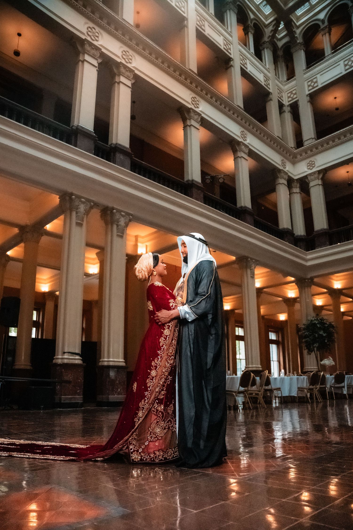Bride and groom in traditional Arabic wedding outfits sharing a romantic moment in a grand hall.