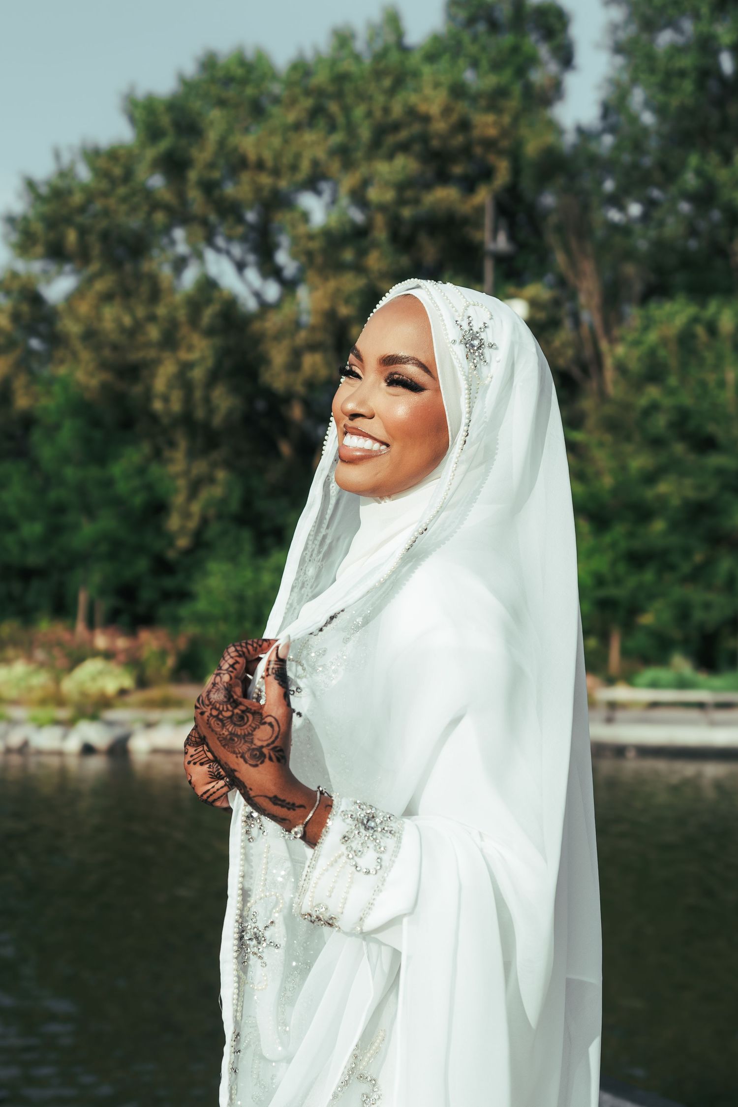Joyful Muslim bride in white hijab wedding dress with intricate henna hands poses by lakeside.
