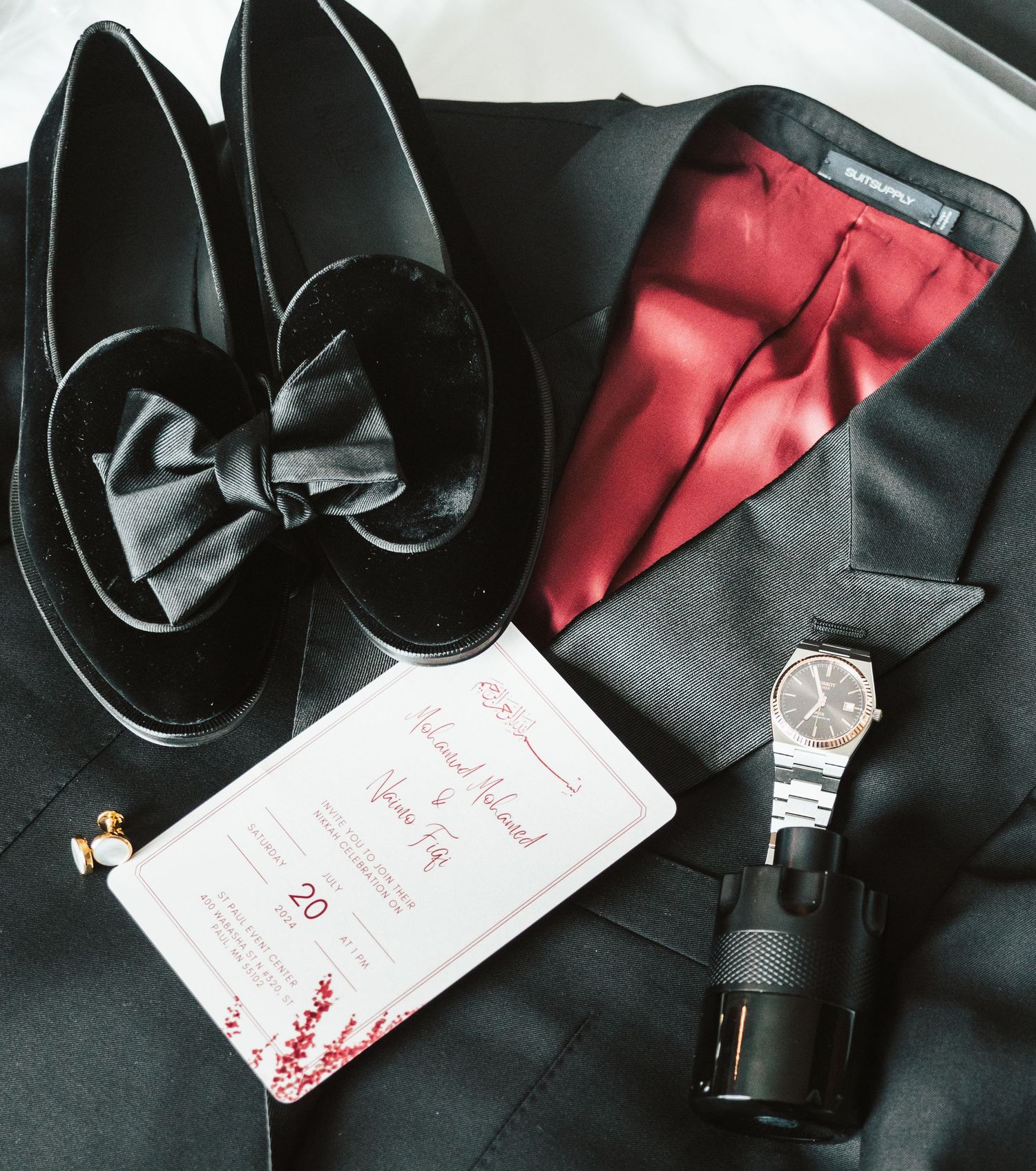Flat lay of black tuxedo, bow tie shoes, watch, cologne, and wedding invitation on suit jacket.
