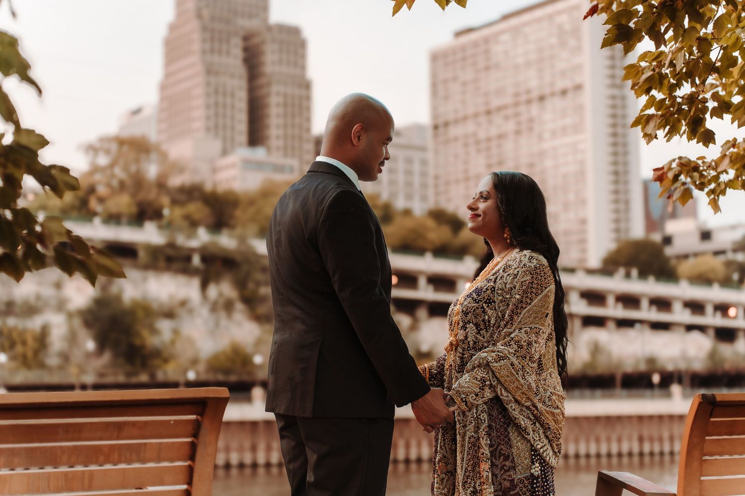 Couple holding hands and gazing at each other with autumn foliage and city skyline in background.