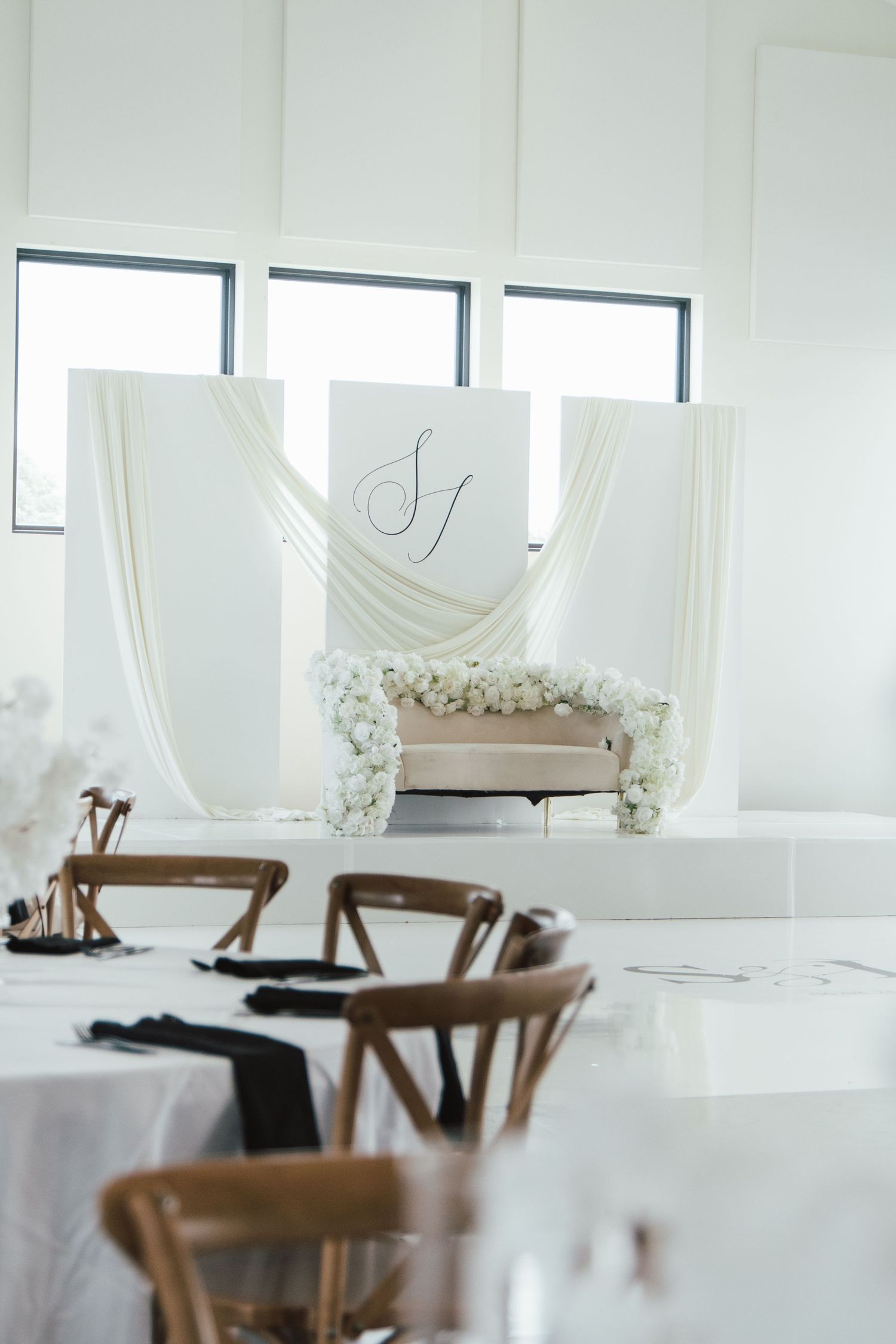 An elegant all-white wedding reception setup with draped fabric, floral arch, and monogram signage.