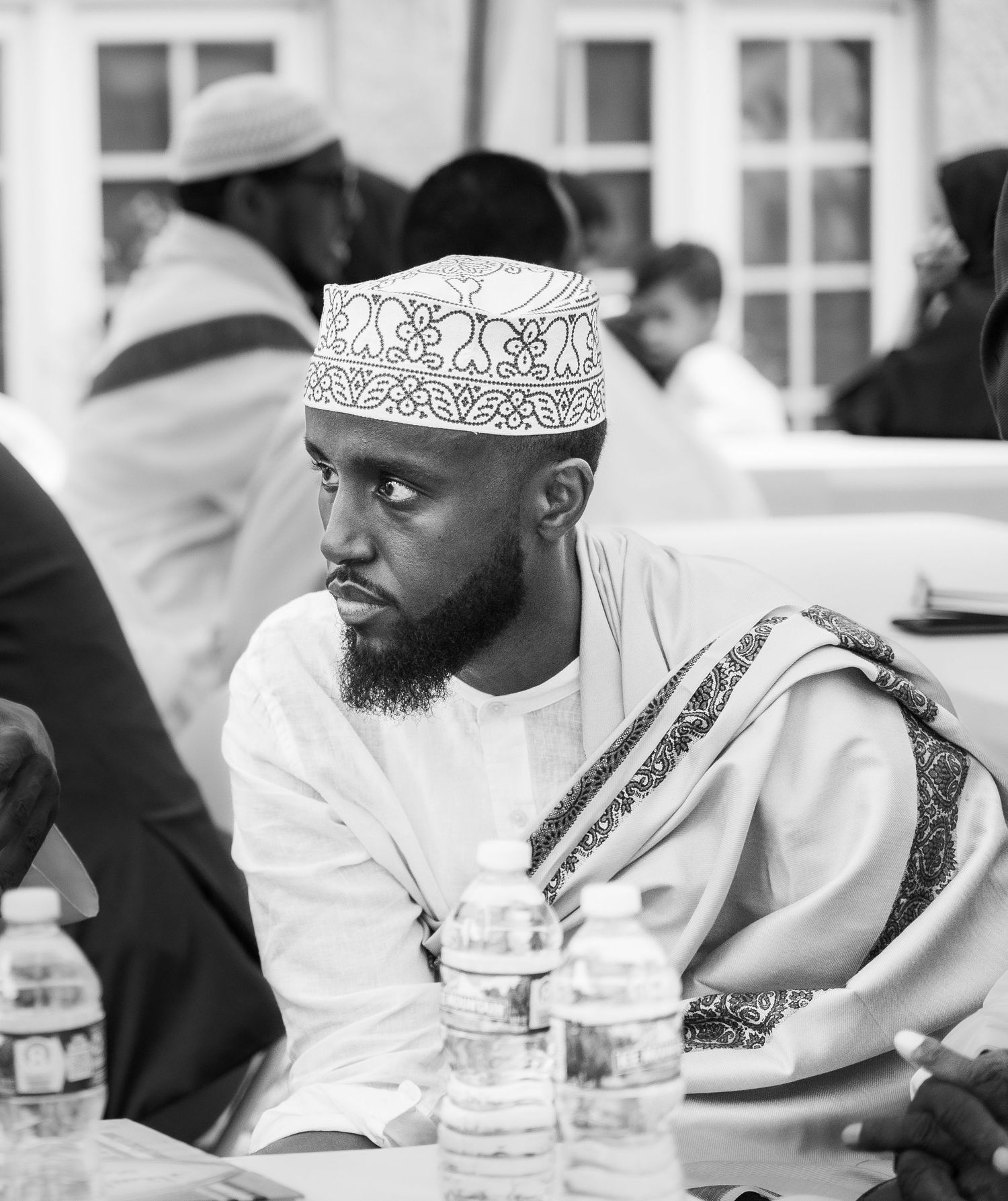 A bearded Muslim man in a white kufi cap and traditional attire sits thoughtfully at an outdoor wedding reception.