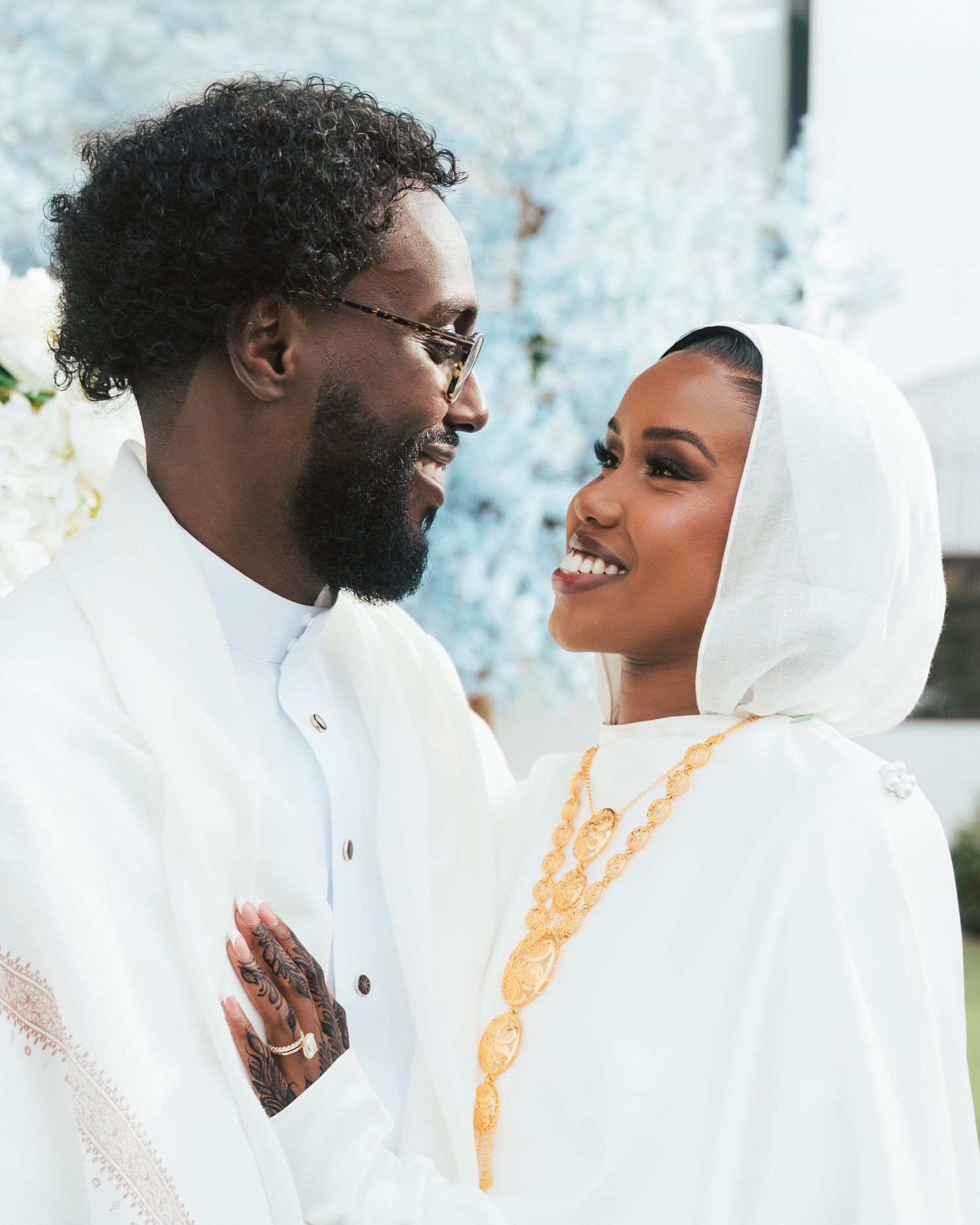 A couple in elegant white traditional attire with gold accessories share a joyful moment at a wedding ceremony.
