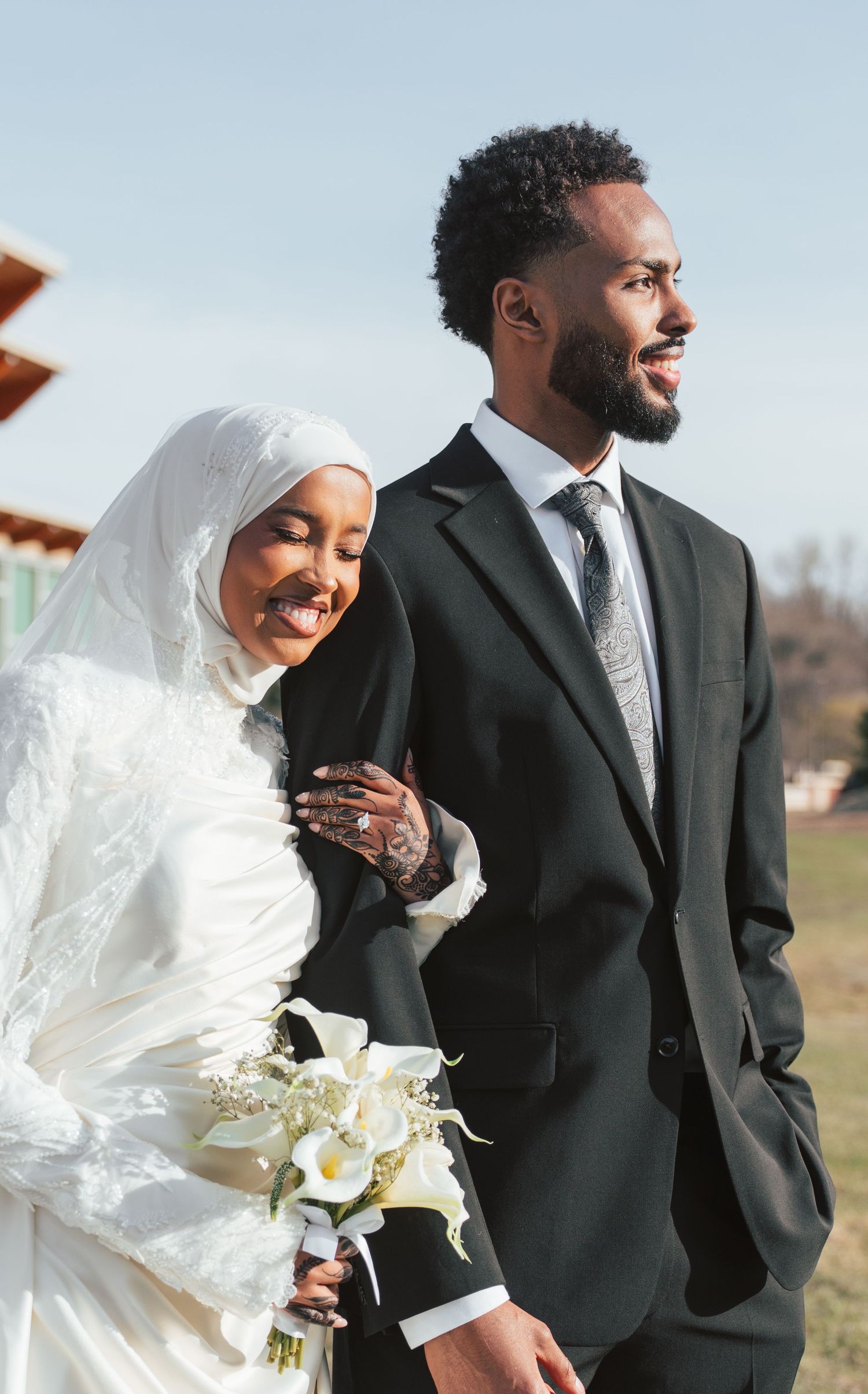Muslim bride in white hijab holding calla lilies bouquet stands with groom in black suit at outdoor wedding.