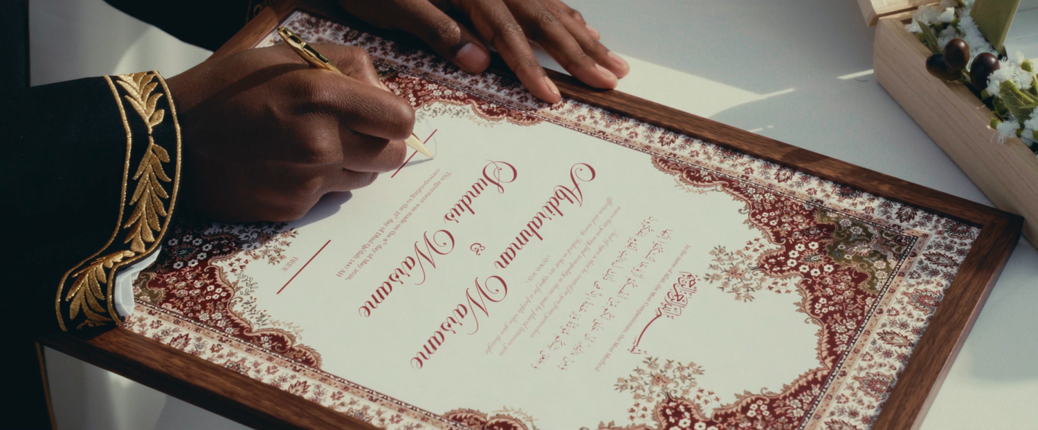 Person signing an ornate wedding certificate with decorative red and gold border design.