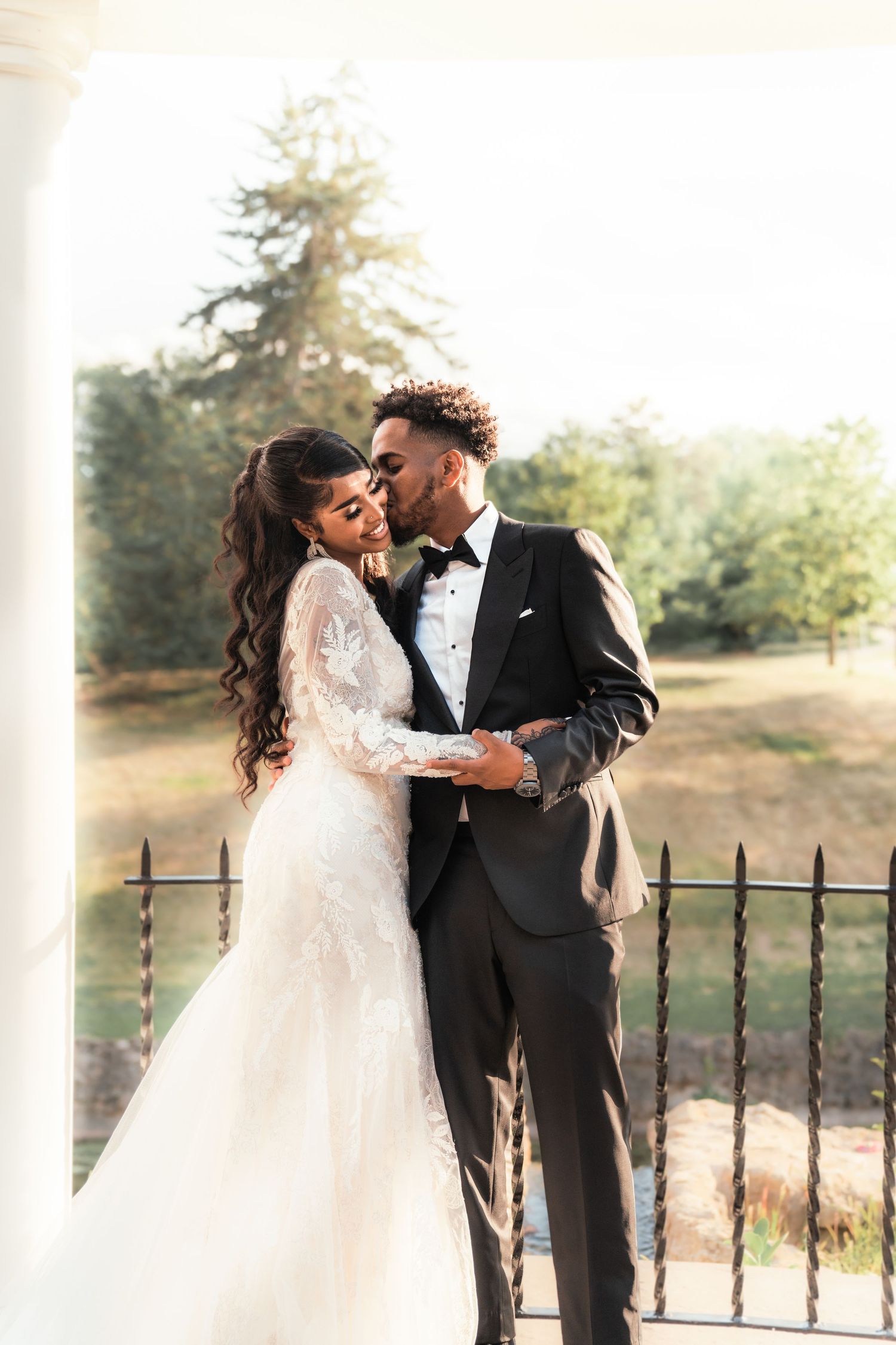 Couple sharing a kiss on a sunlit porch, bride in lace gown and groom in black tuxedo.
