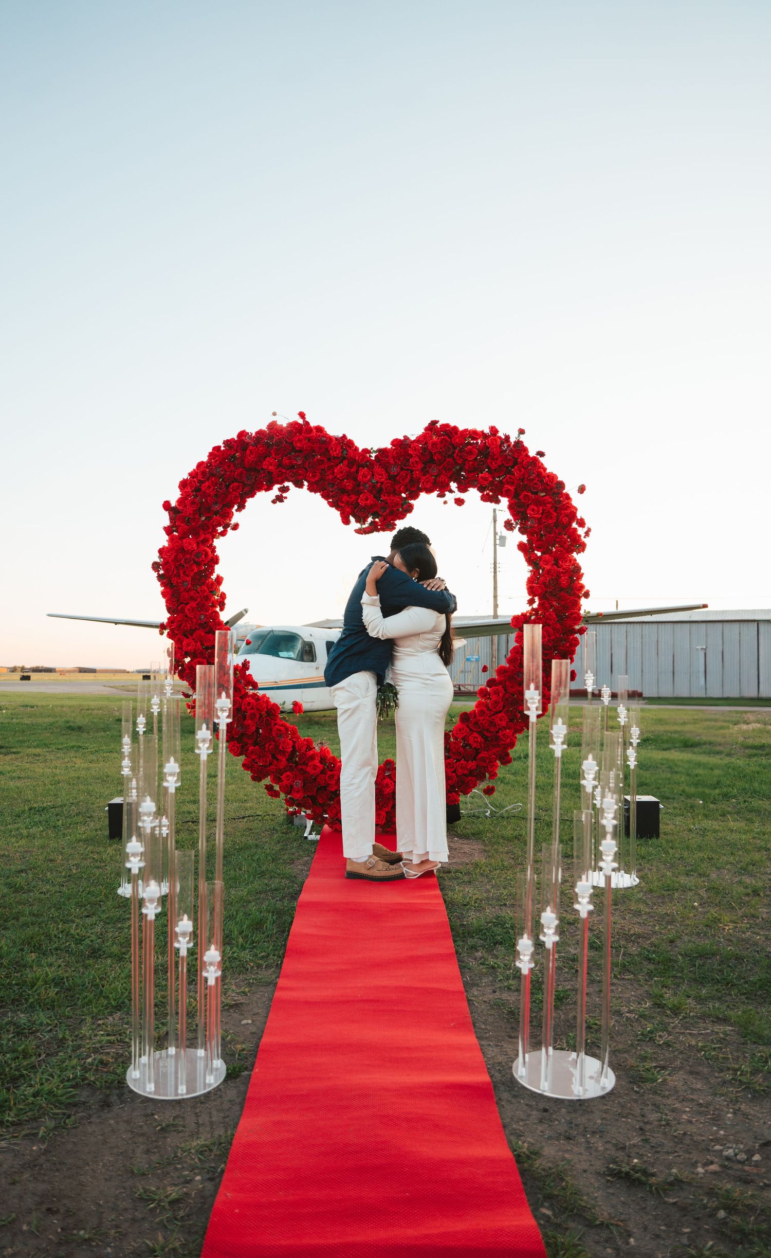 Couple embracing under large red rose heart arch on red carpet at romantic outdoor airport proposal setup.