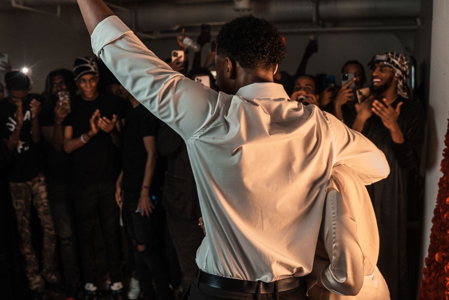 Groom in white shirt celebrates with raised arm surrounded by cheering guests at wedding reception.