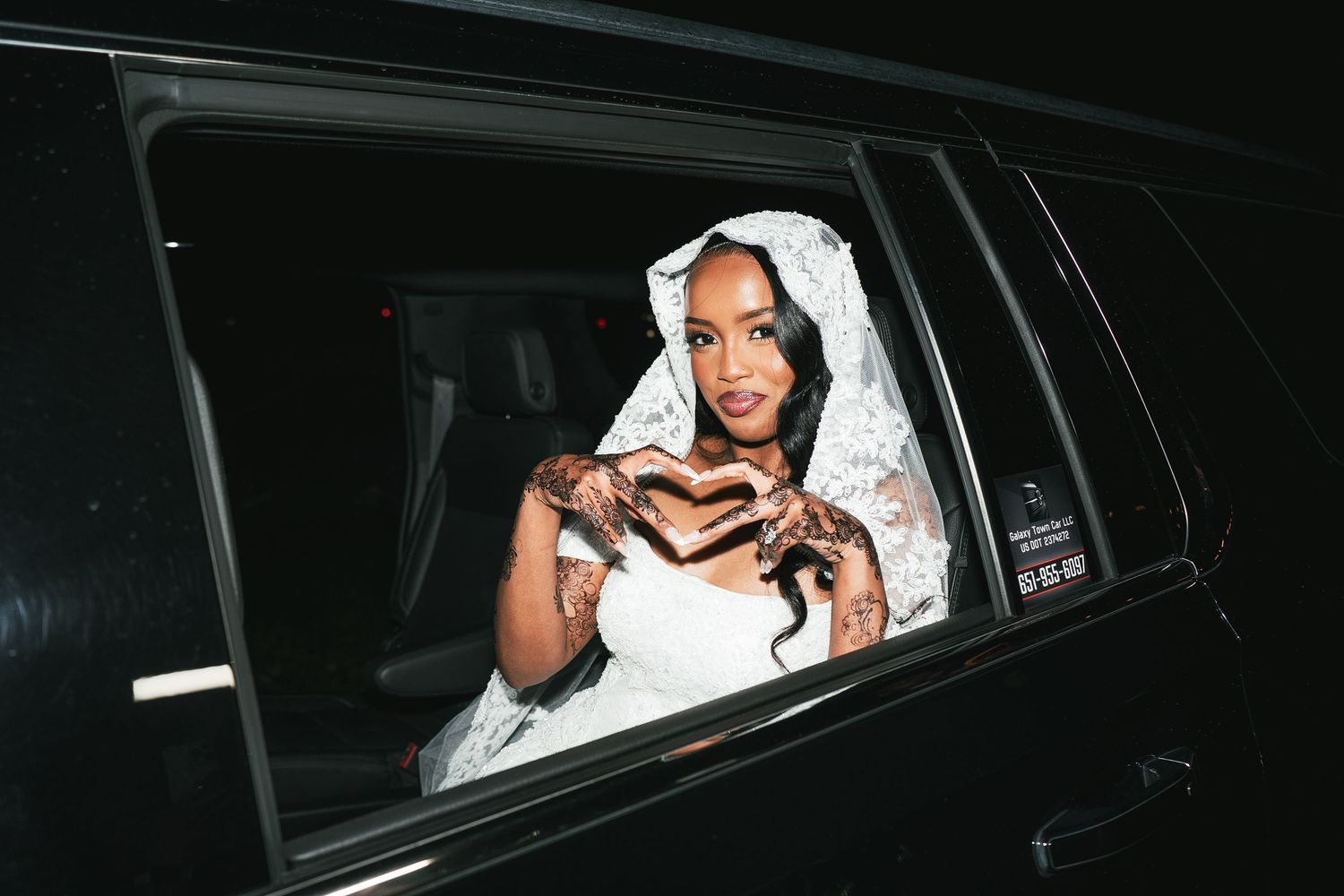 Woman in white lace bridal gown and veil making heart shape gesture inside a luxury black vehicle at night.