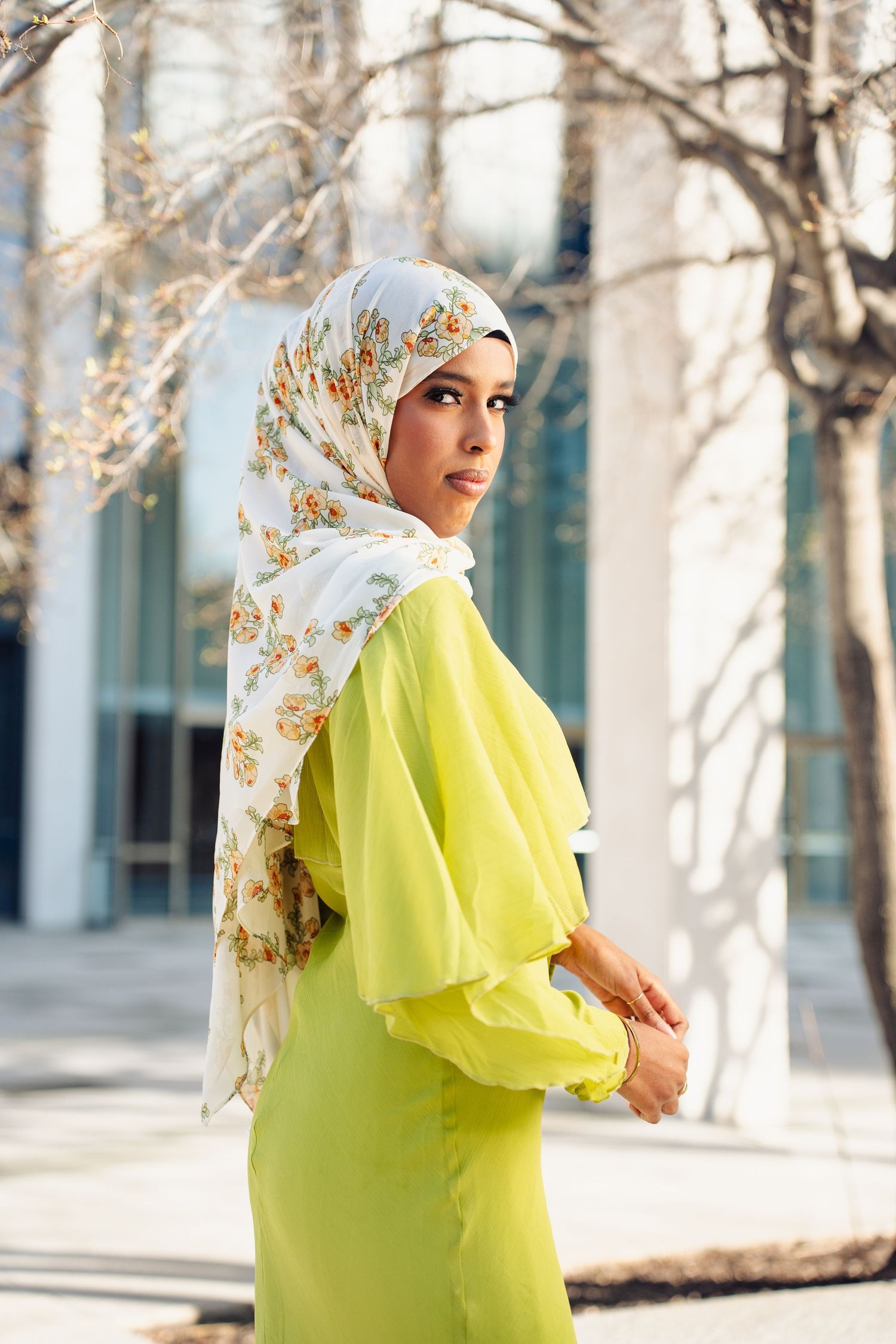 Stylish woman in bright yellow-green outfit wearing floral hijab posing outdoors near modern glass building.