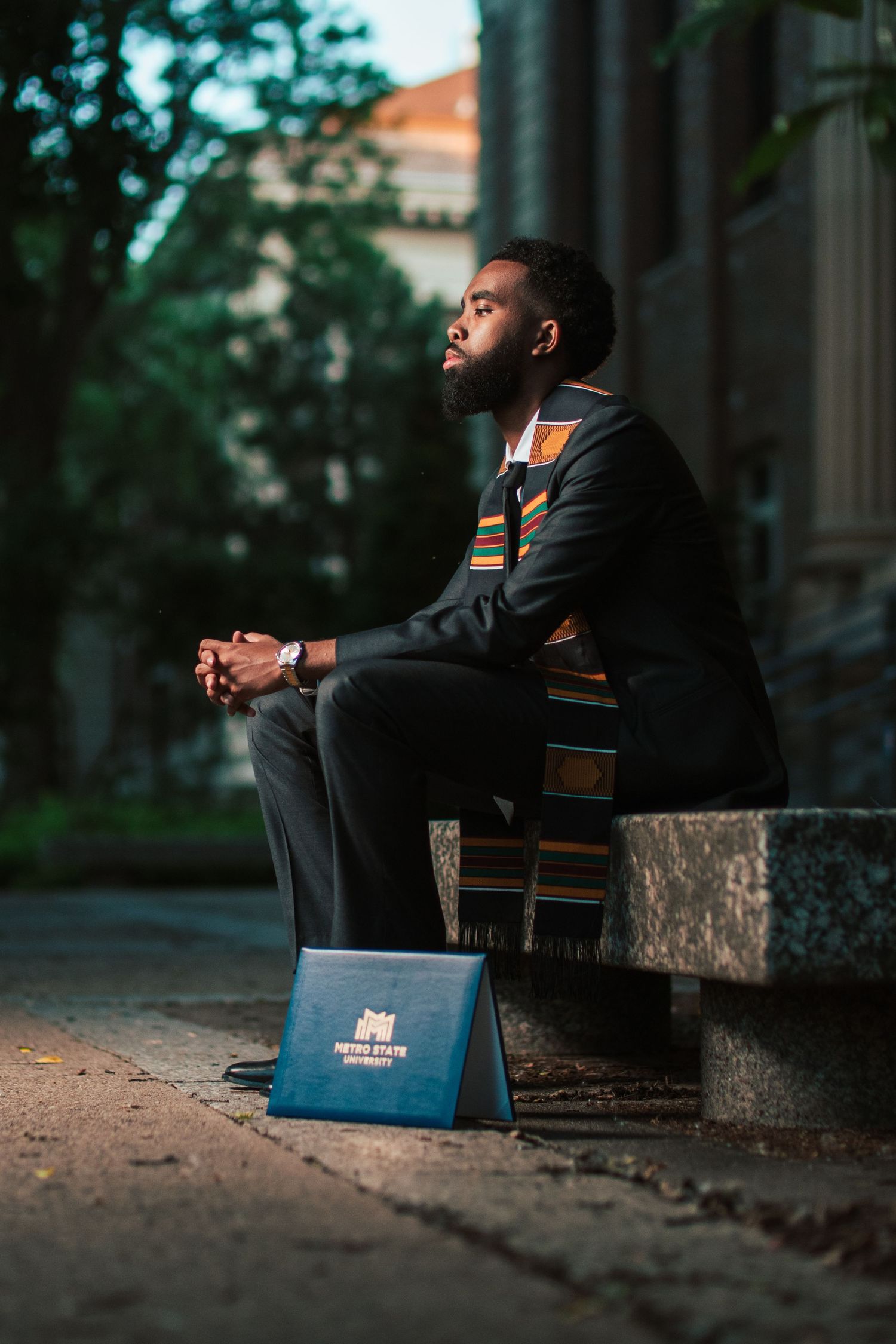 Black male graduate in suit and kente stole sits pensively on stone steps with diploma folder.