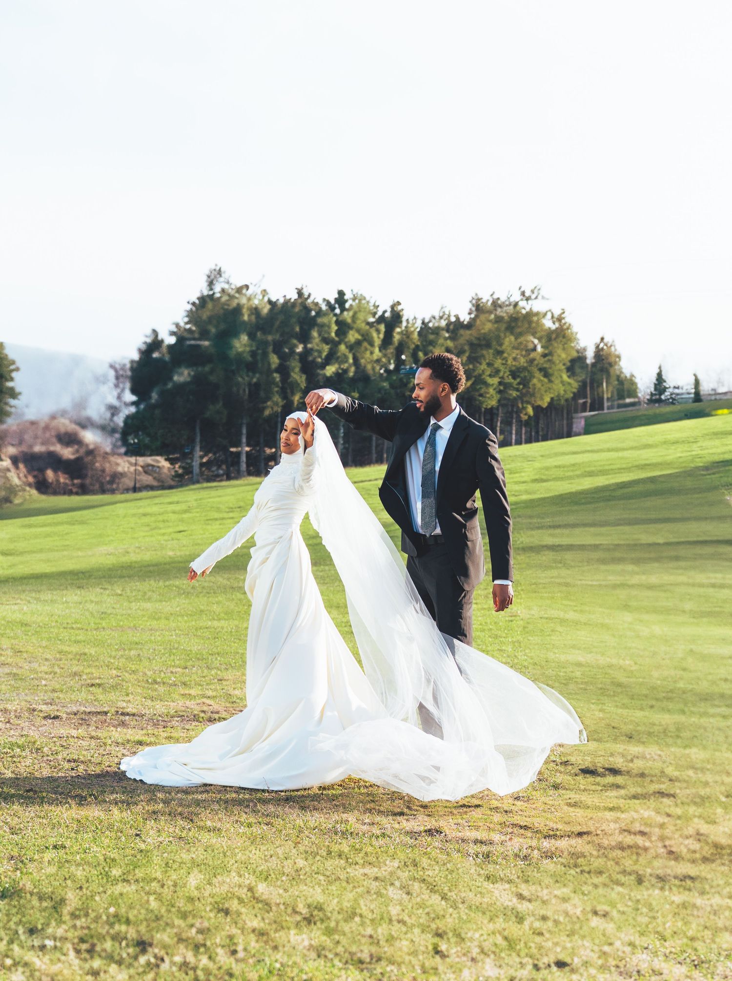 Groom in black suit twirls Muslim bride in flowing white hijab dress on green lawn at wedding.