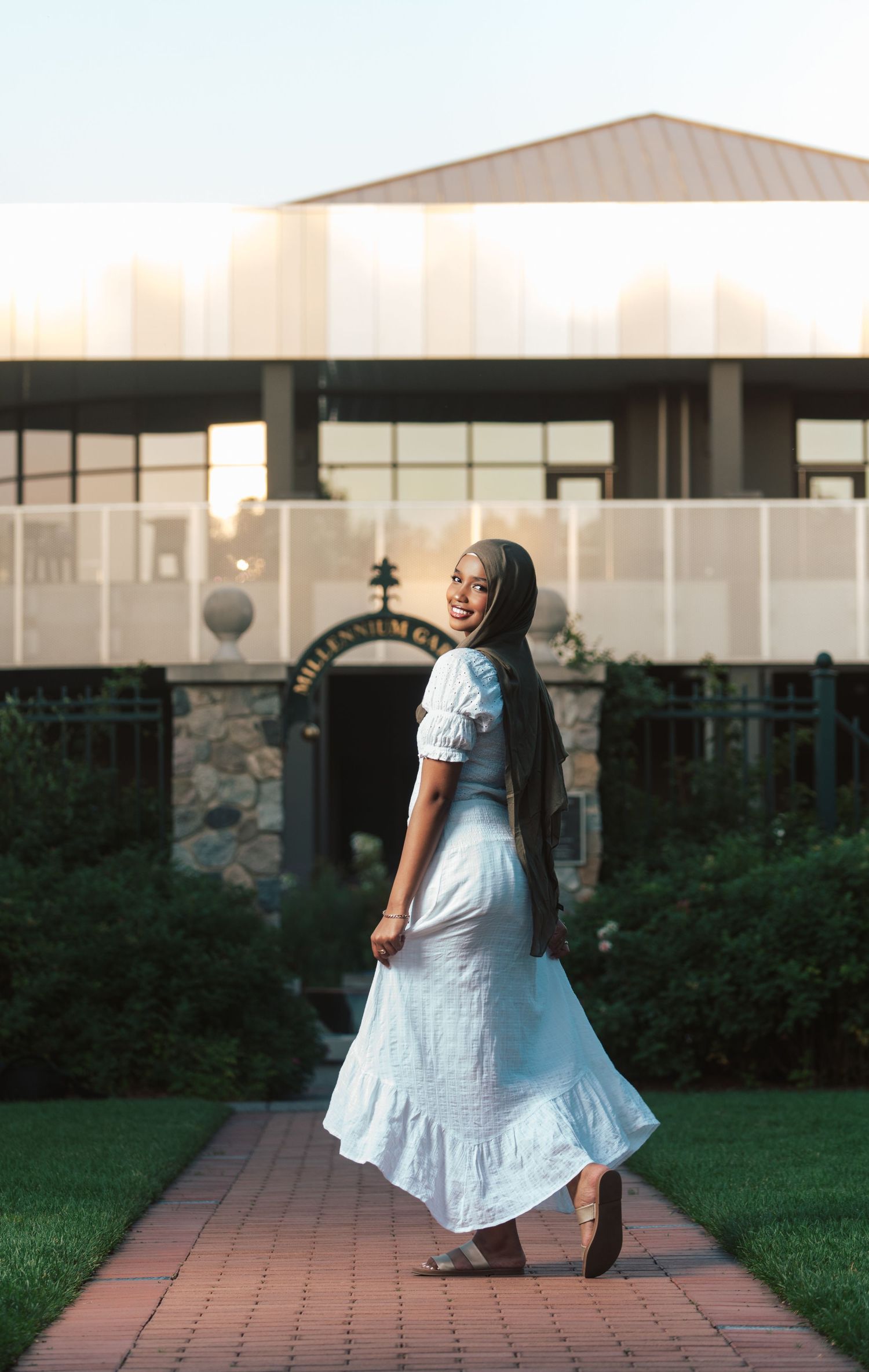 Woman in white midi dress and hijab smiles outside Millennium Garden at golden hour.