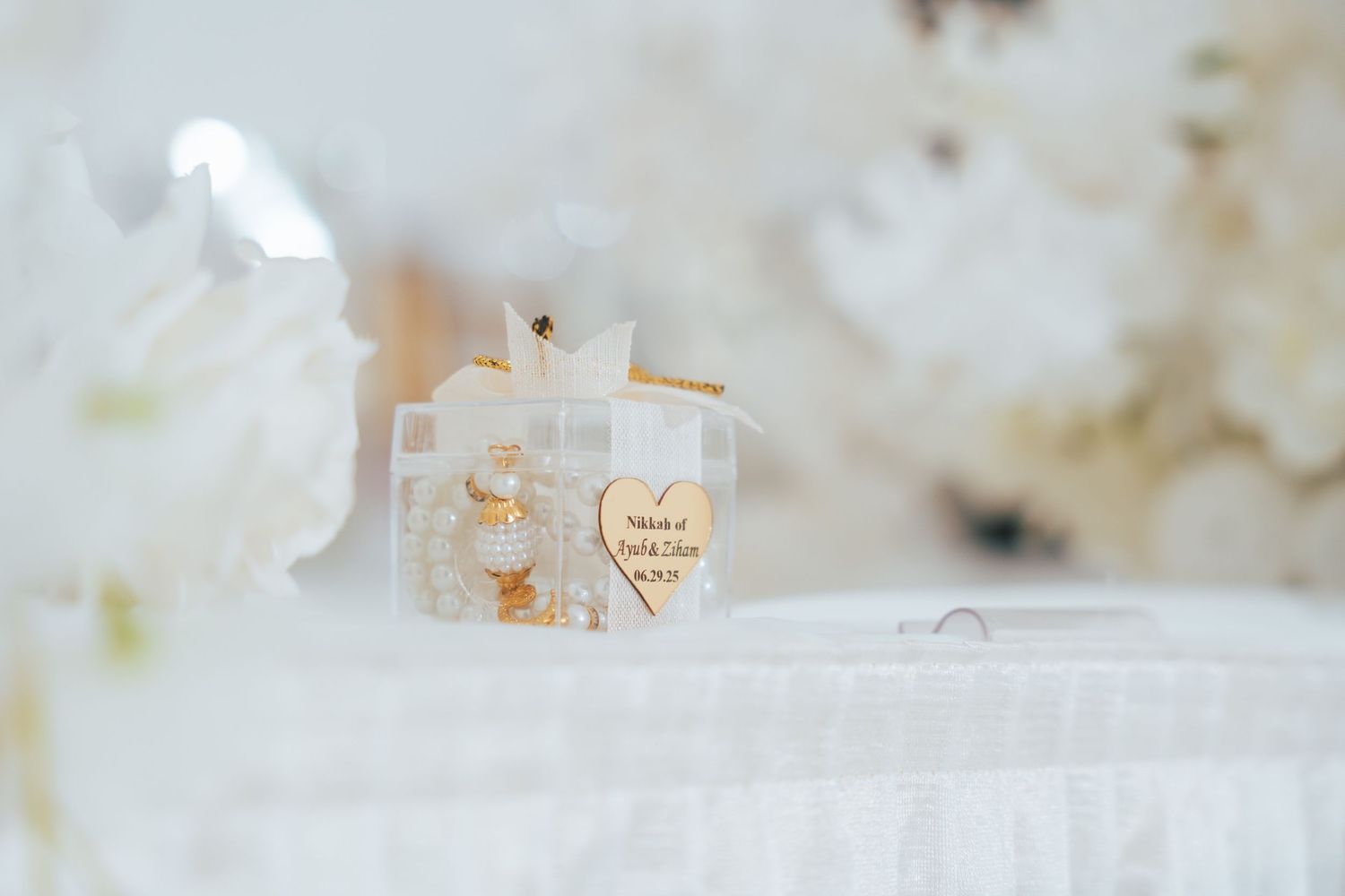 A decorative personalized wedding favor box with gold heart charm surrounded by white floral arrangements.