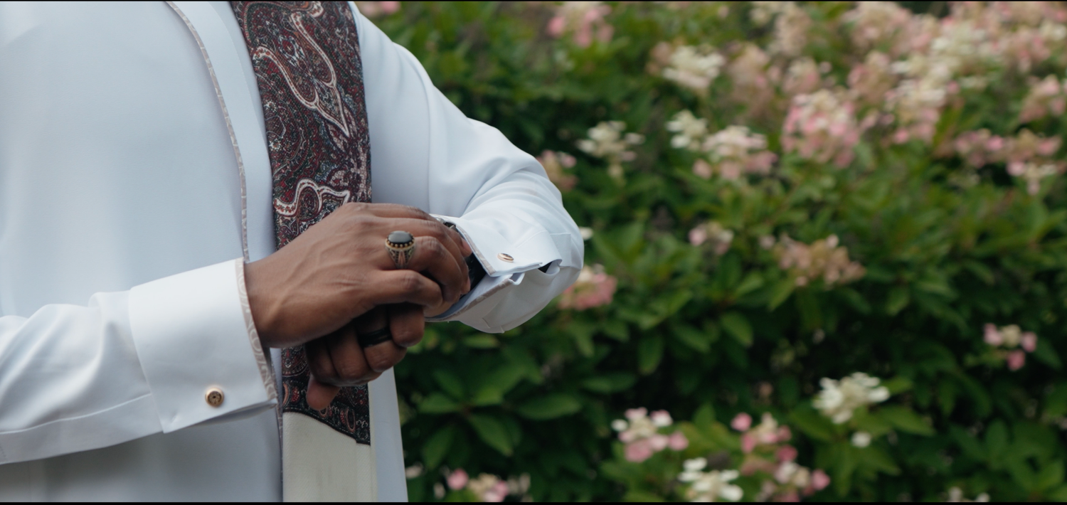 Man in white dress shirt adjusting cufflinks with pink floral bushes in background.