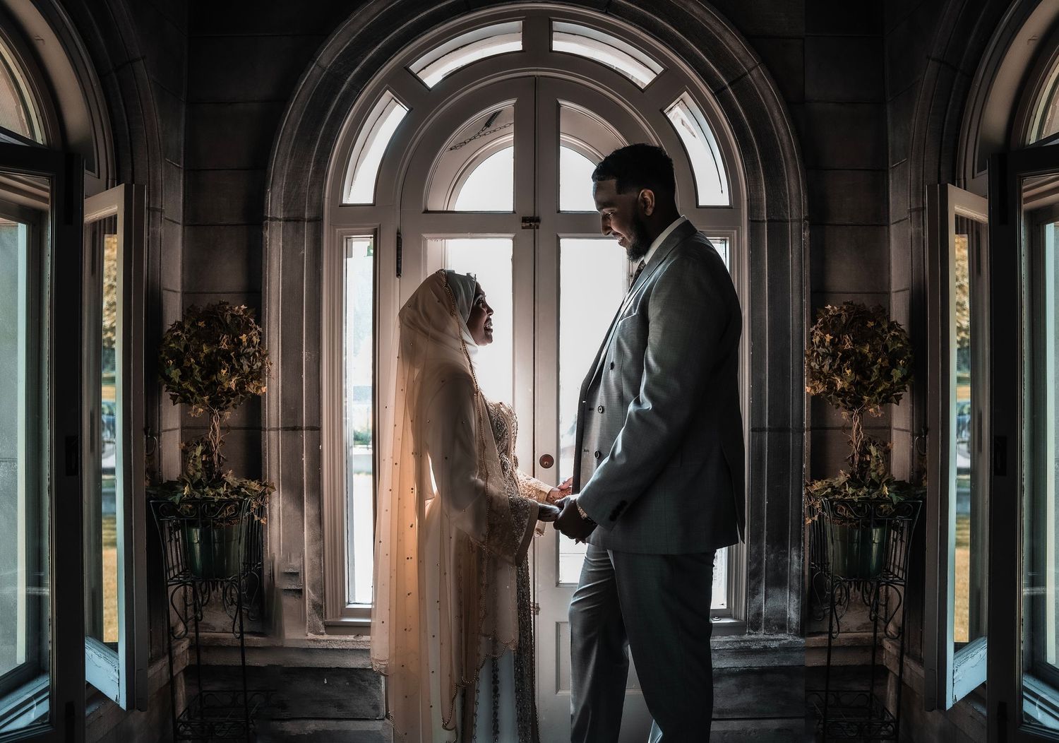 A Muslim bride in hijab and groom in a grey suit hold hands by an arched window in an elegant venue.