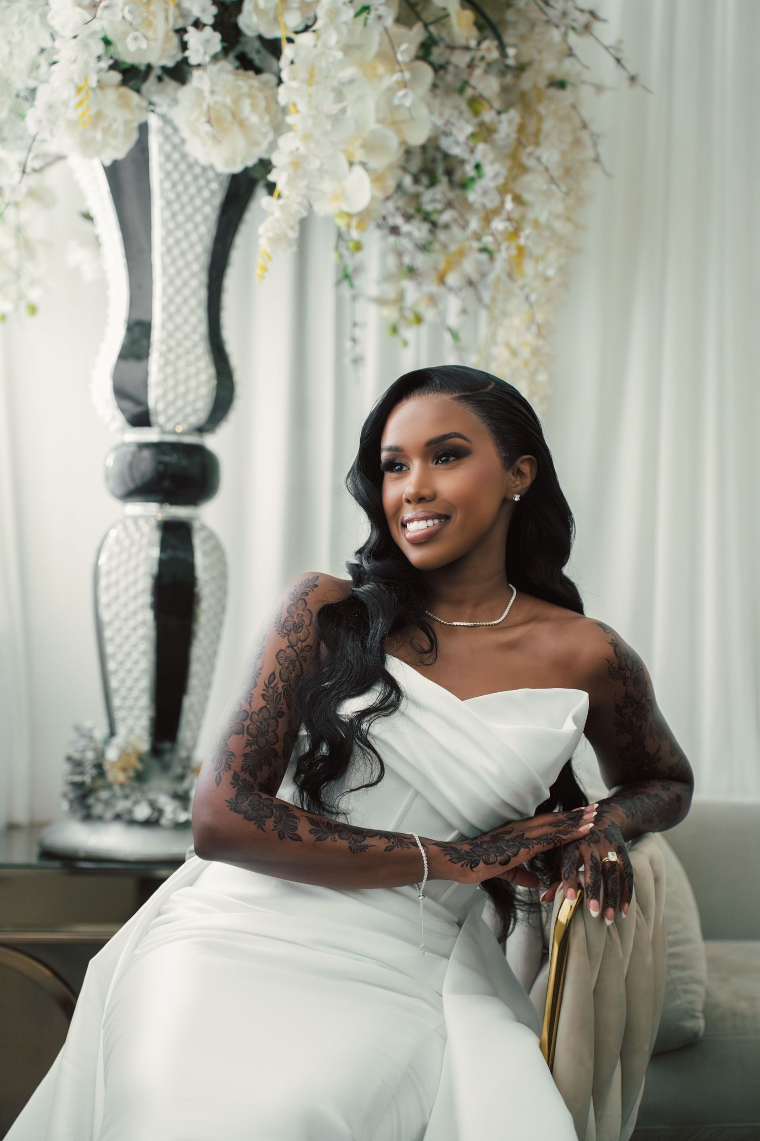 Smiling bride in white strapless gown with tattoos sits before white floral arrangement in elegant wedding setting.