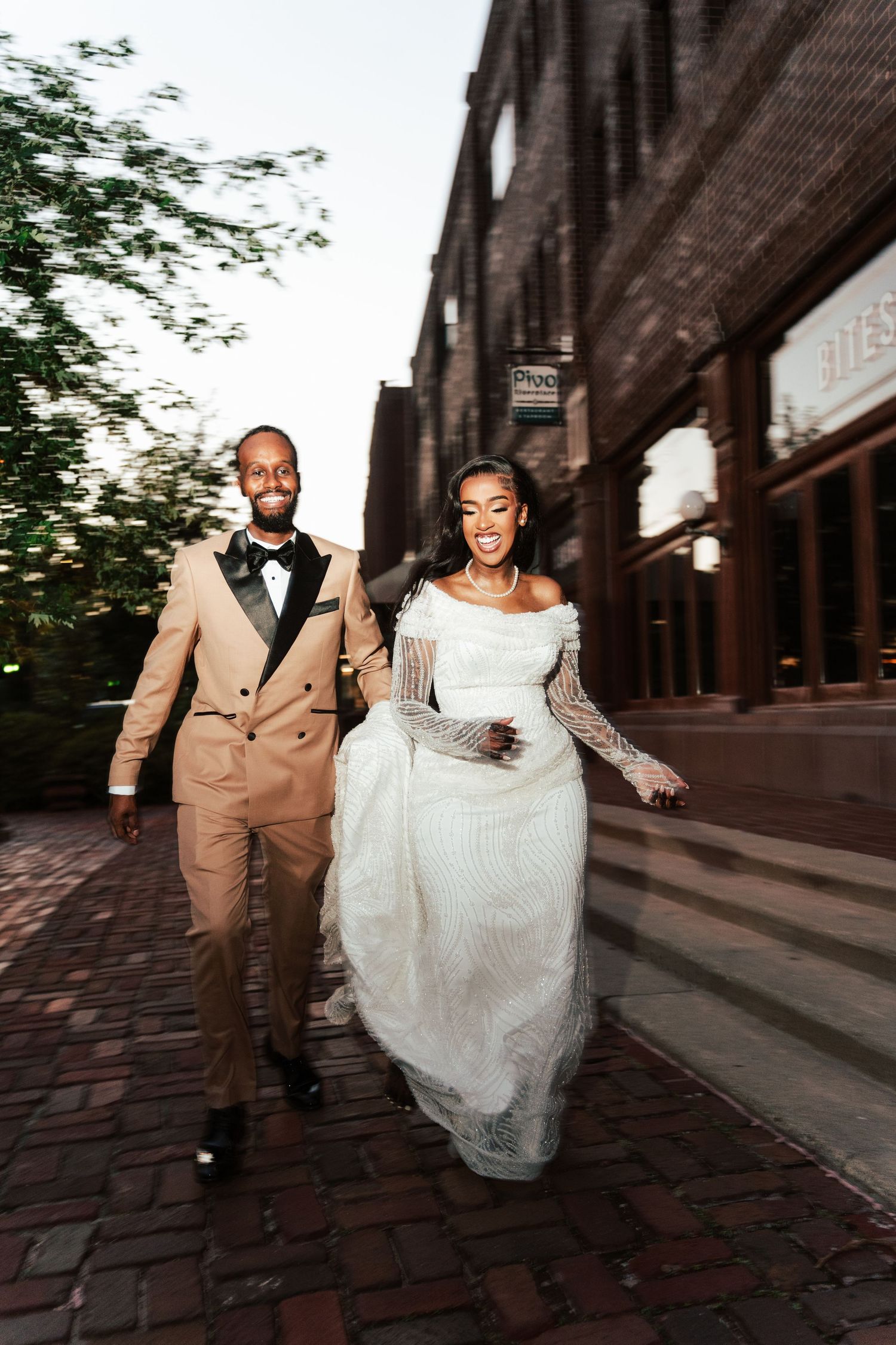 A joyful Black bride and groom in elegant wedding attire walk hand in hand on a brick city street.