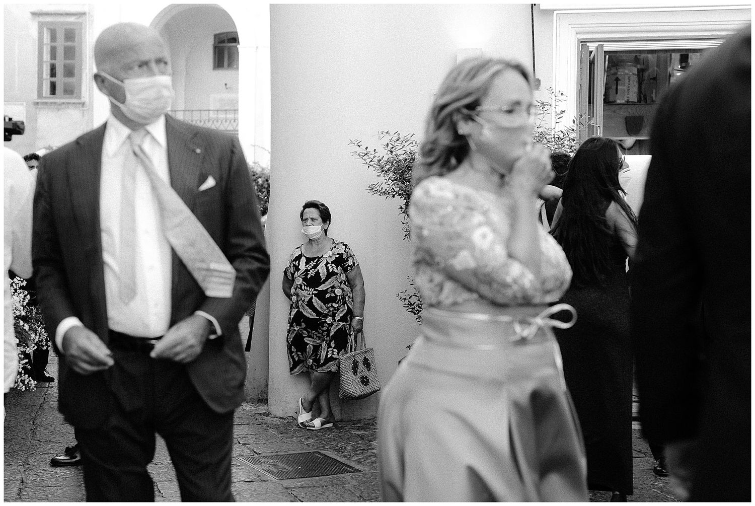 Black and white photo of wedding guests mingling outside in formal attire with someone in white dress moving through the crowd.