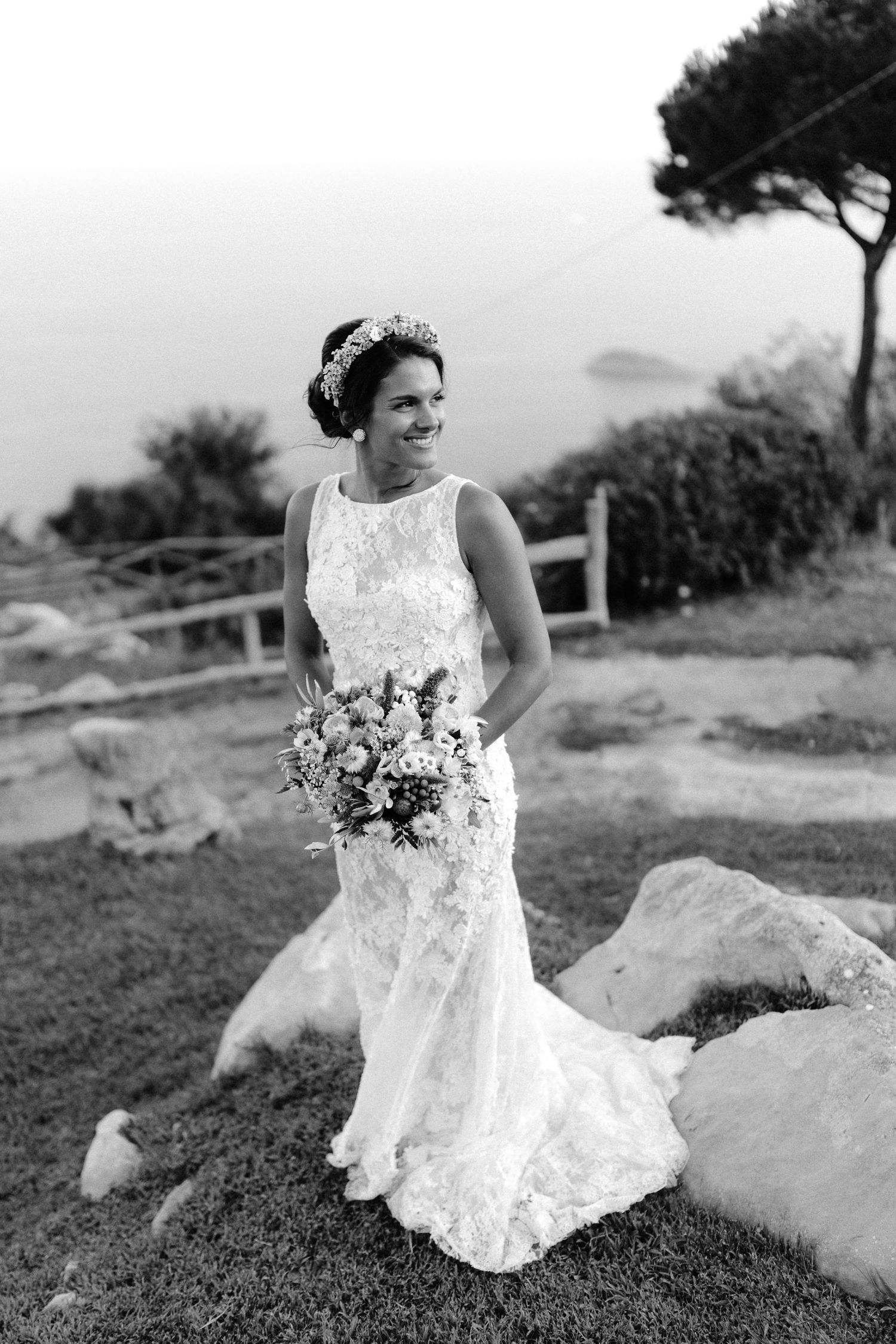 Elegant bride in a fitted lace wedding gown holding a floral bouquet poses outdoors against a coastal landscape.