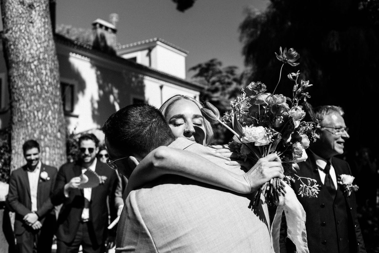 A black and white candid shot captures a warm embrace at a wedding ceremony with onlookers and flowers visible.