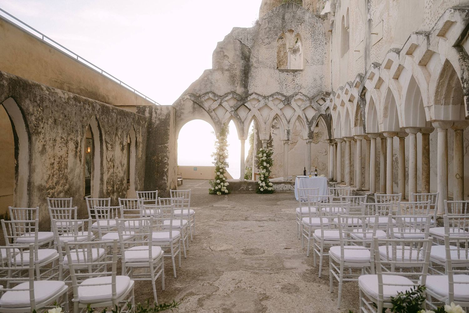 White chairs arranged in rows at an elegant stone cloistered venue with arched windows for a wedding ceremony.