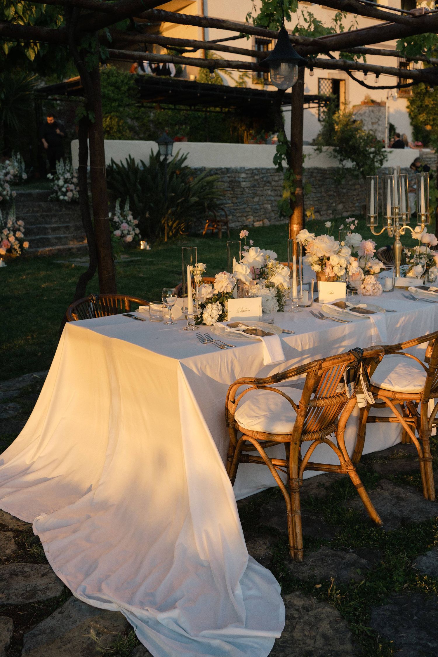 Elegant outdoor wedding reception table with white linen, floral centerpieces, candles, and rattan chairs at dusk.