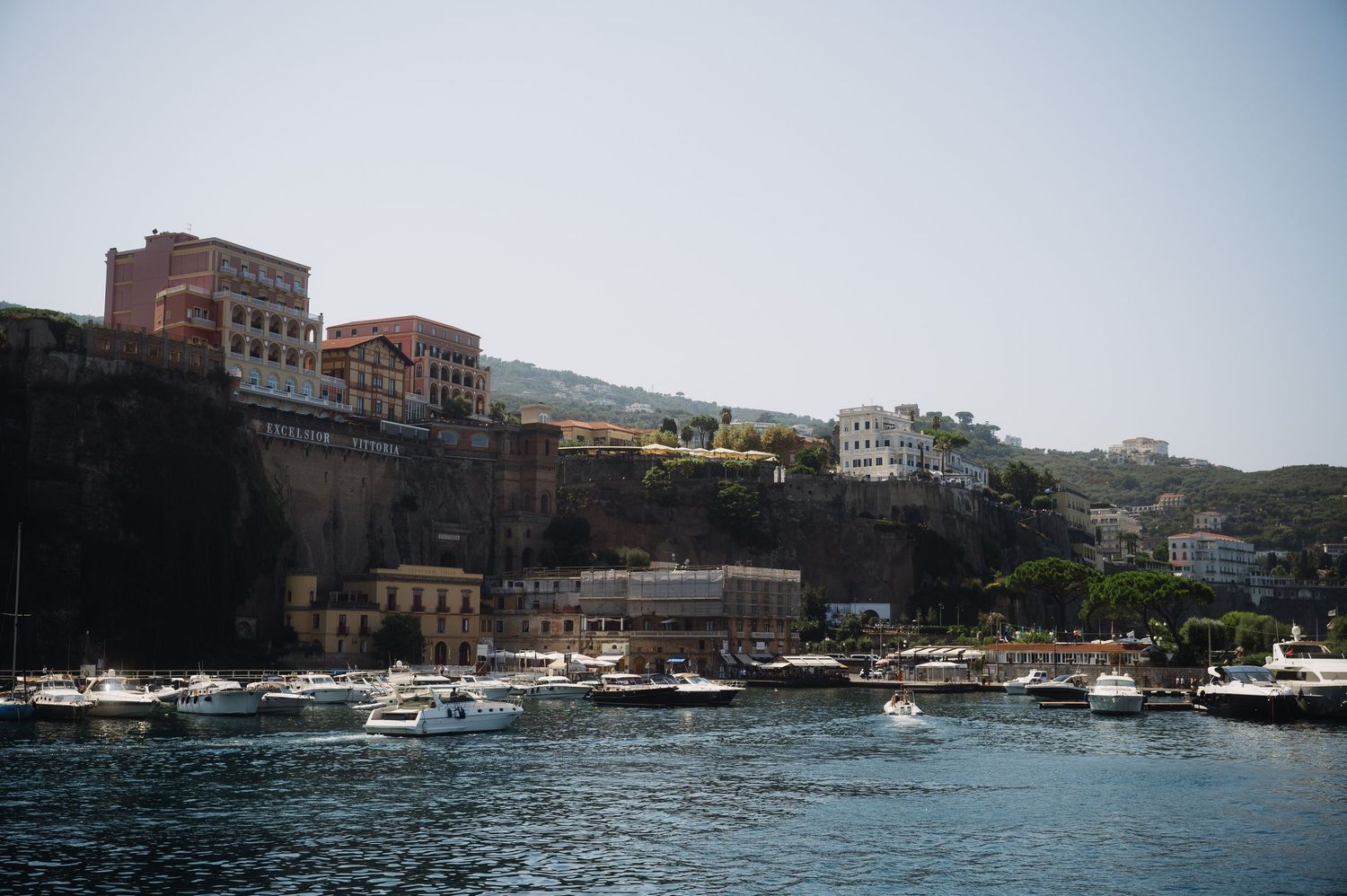 Panoramic view of boats docked in a Mediterranean harbor with historic buildings along the waterfront.