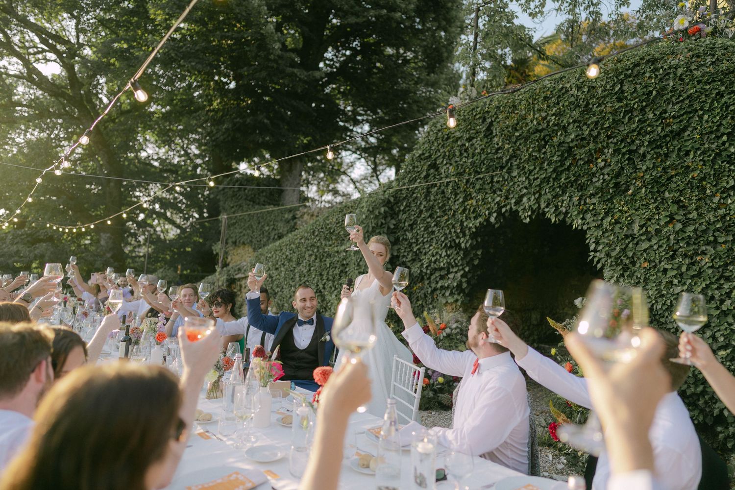 Guests at an outdoor wedding reception raise their hands in celebration under string lights and trees.