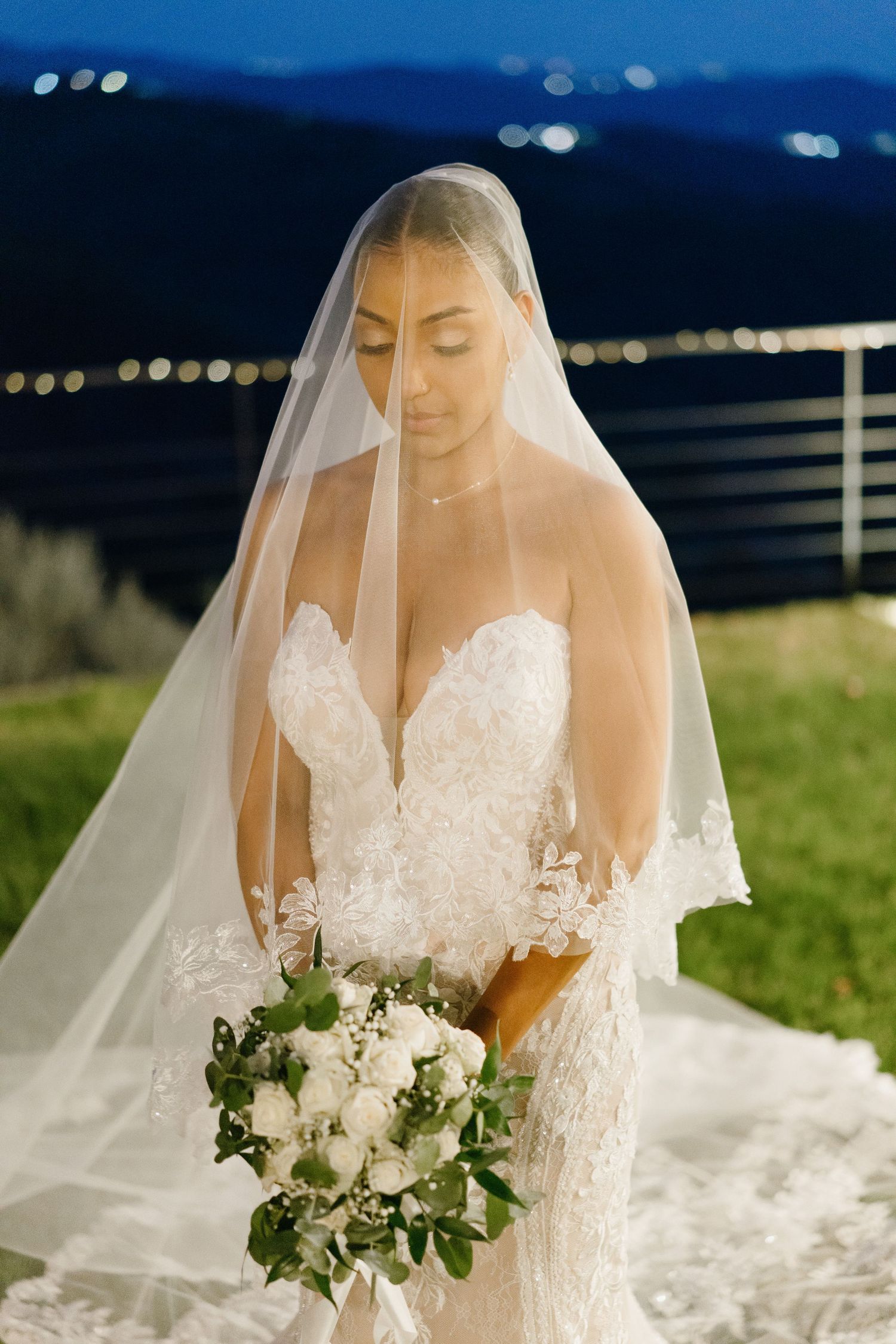 A bride in a white lace wedding dress and veil holds an elegant white floral bouquet during an outdoor ceremony.