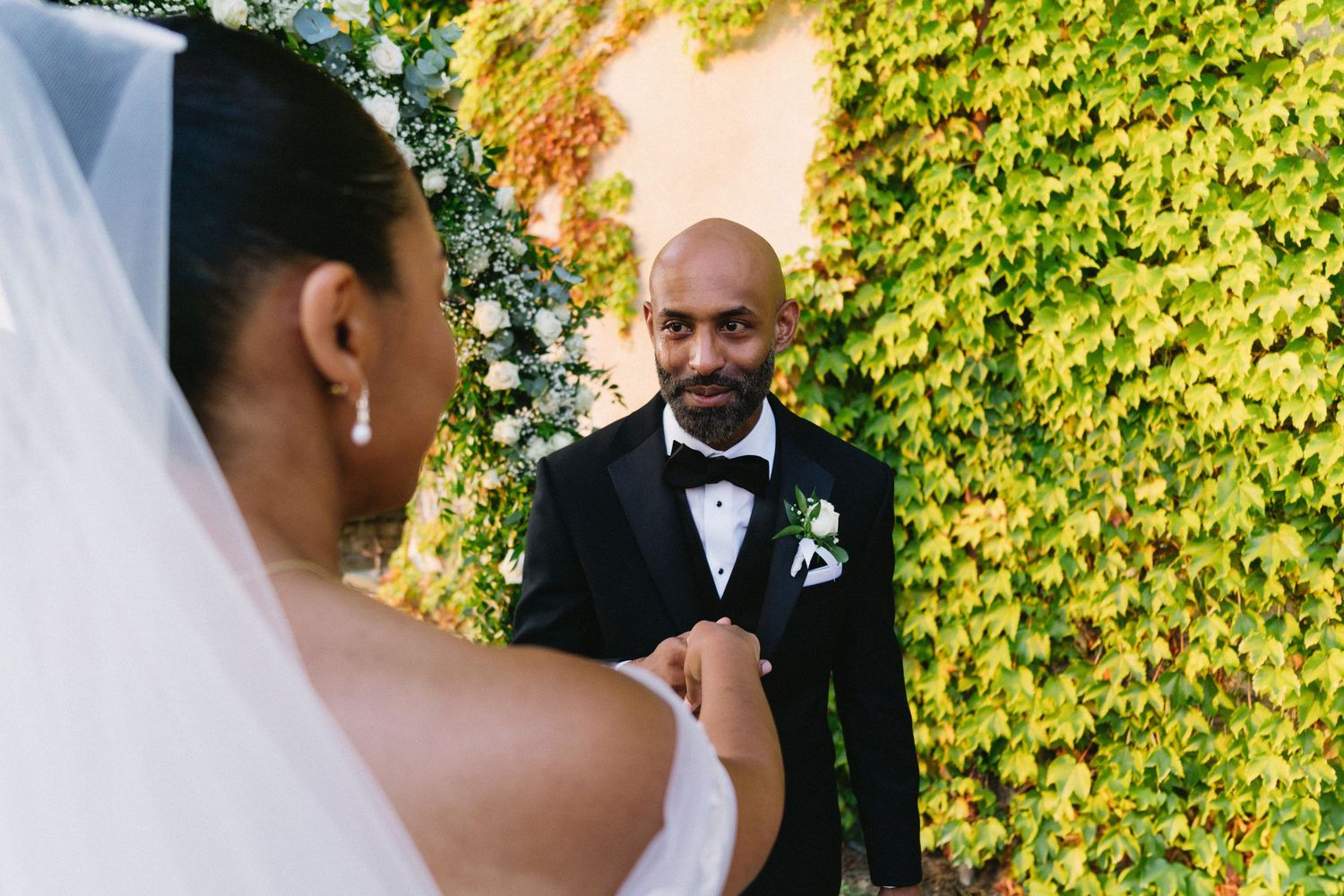 A wedding ceremony takes place outdoors with lush green ivy-covered walls creating a natural backdrop.