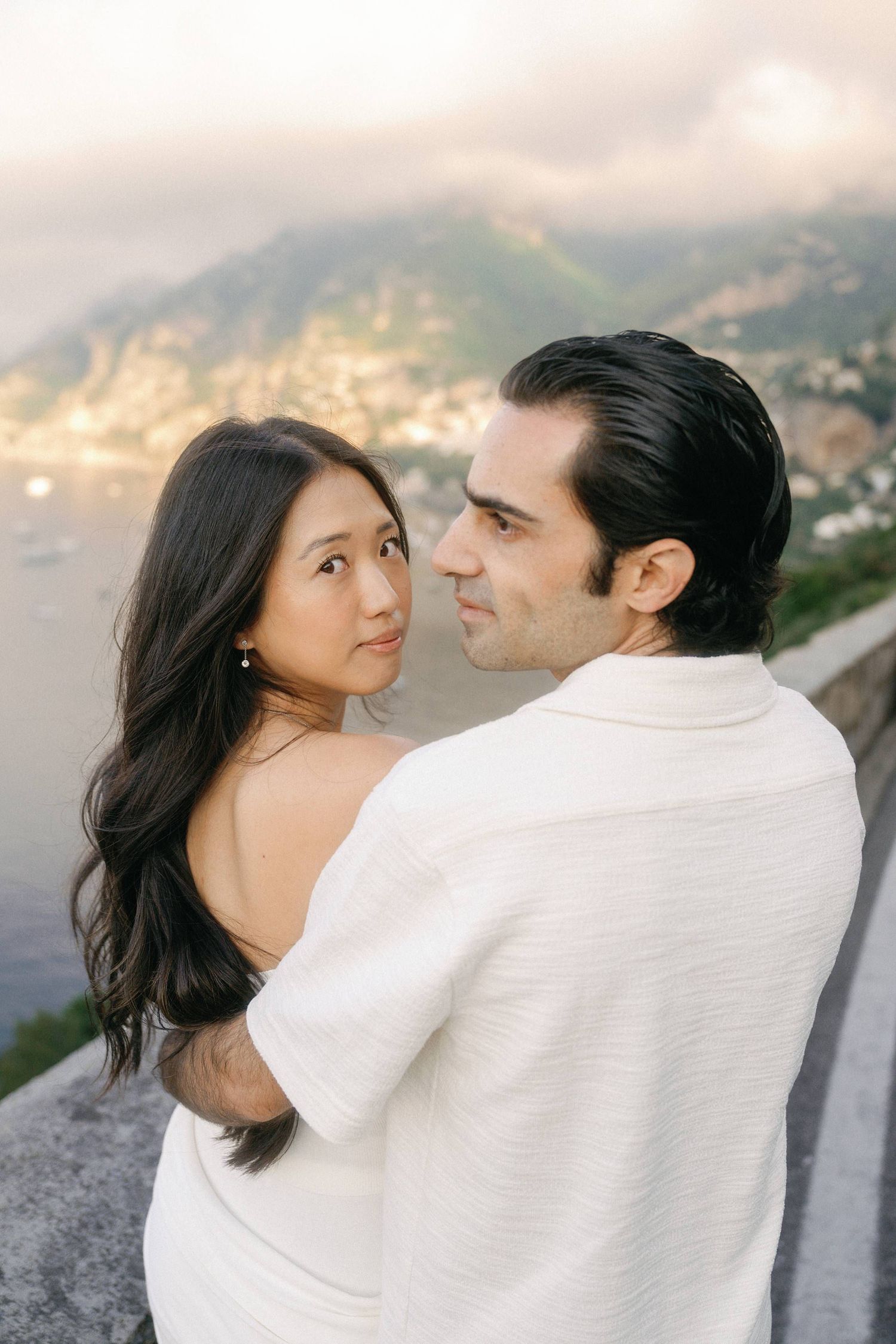 Romantic couple embraces against scenic coastal mountain backdrop during golden hour.