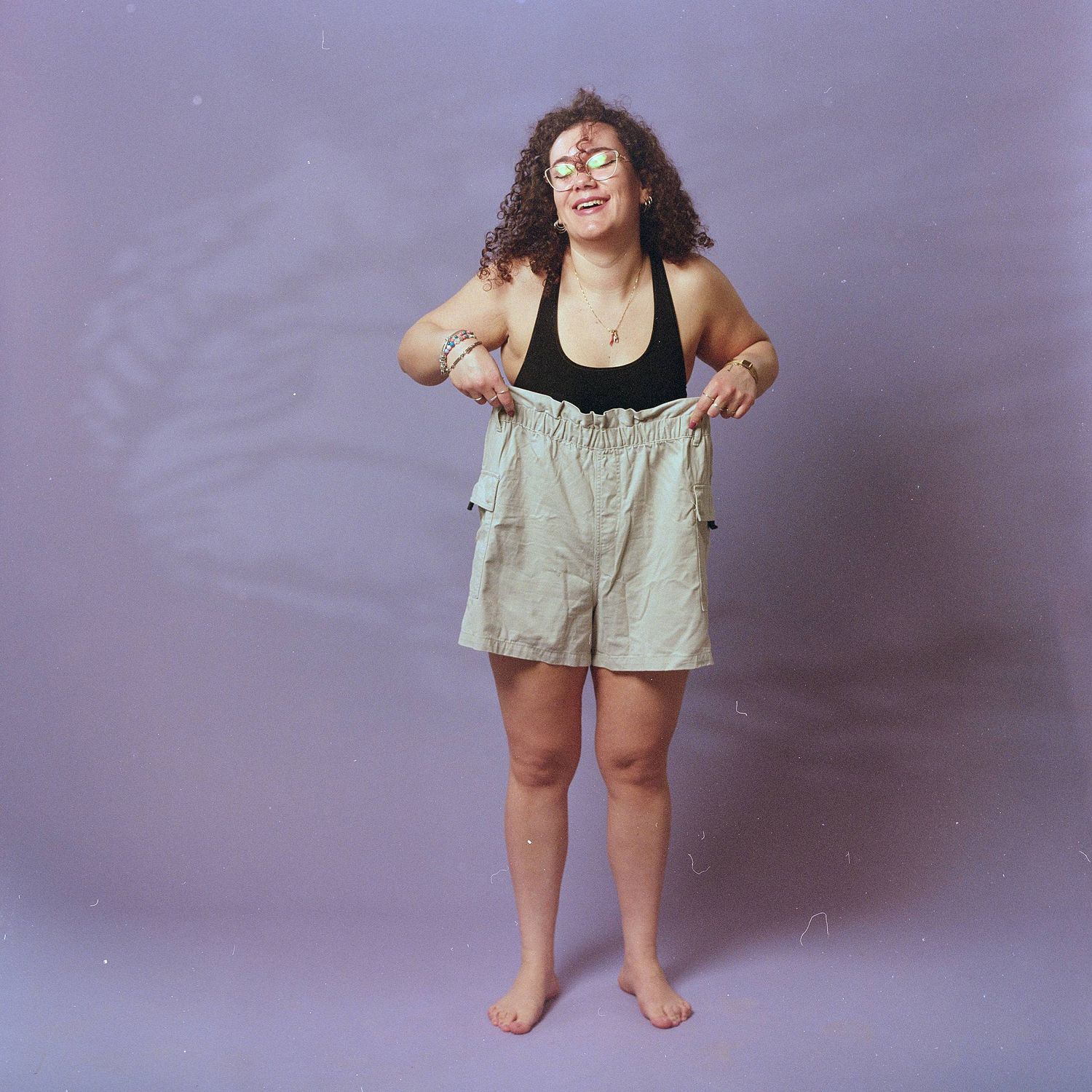 A person with curly hair wearing black tank top and beige shorts stands barefoot against purple backdrop while smiling.