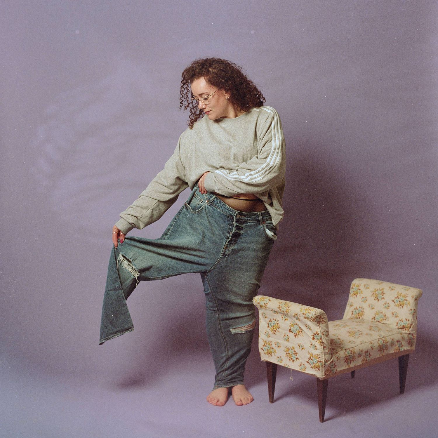 A barefoot person in ripped jeans and cream sweater poses artistically next to a vintage floral upholstered chair against purple wall.