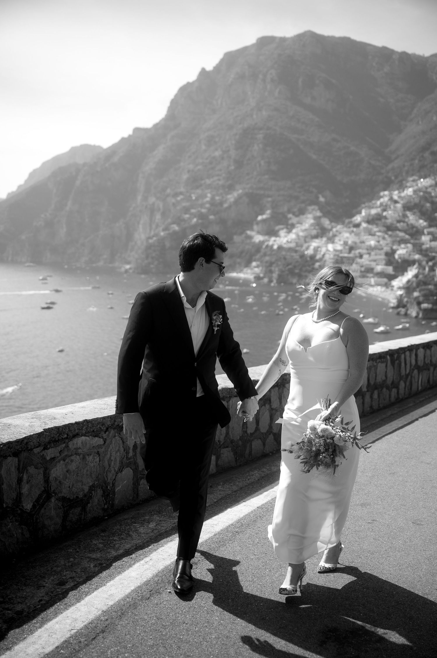 A couple in wedding attire walk along a scenic coastal road overlooking cliffs and Mediterranean waters in black and white.