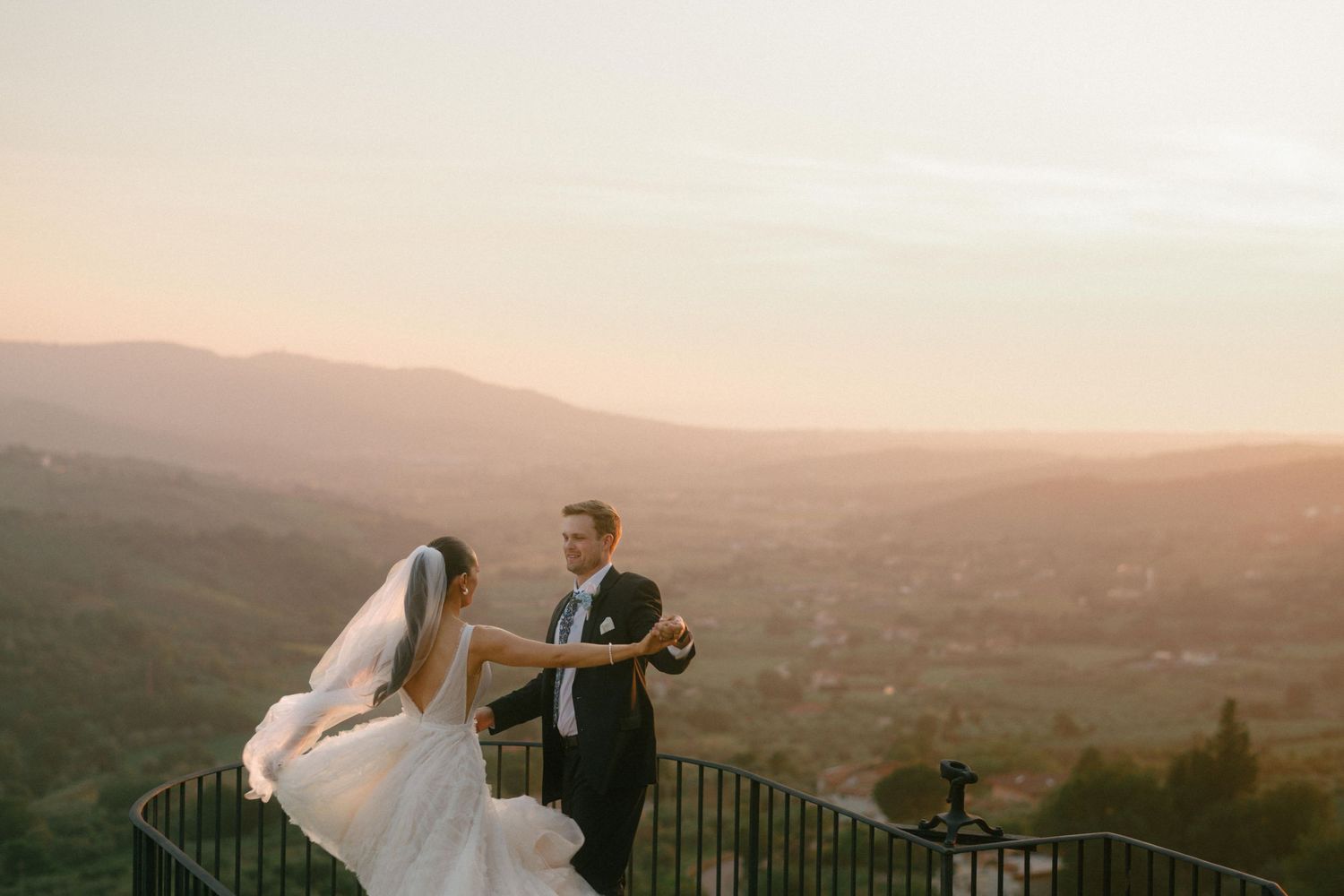 A couple in wedding attire shares a dance on a hilltop terrace overlooking a scenic mountain landscape at sunset.