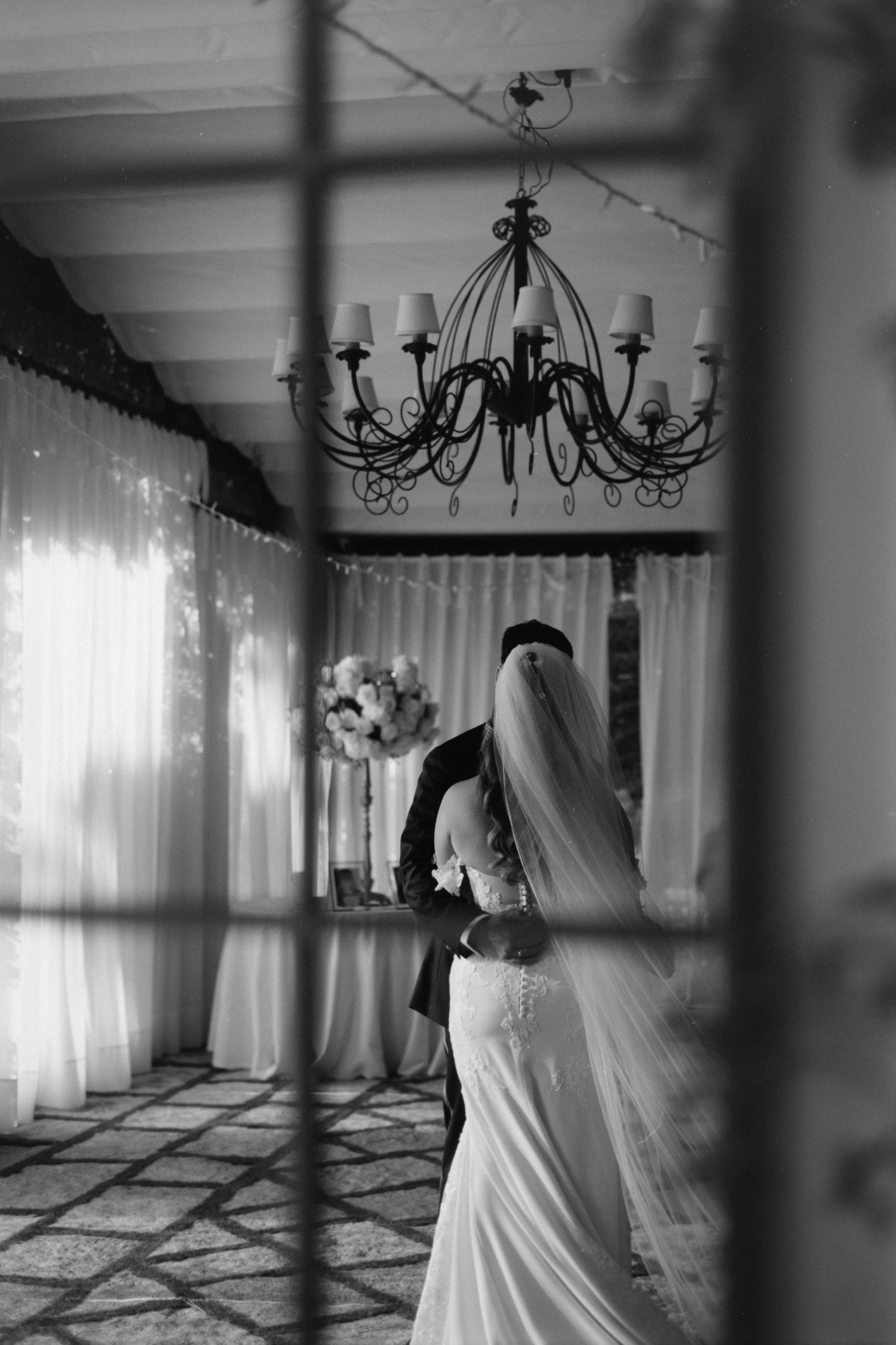 Black and white wedding photo showing a bride in an elegant dress beneath a dramatic wrought iron chandelier.