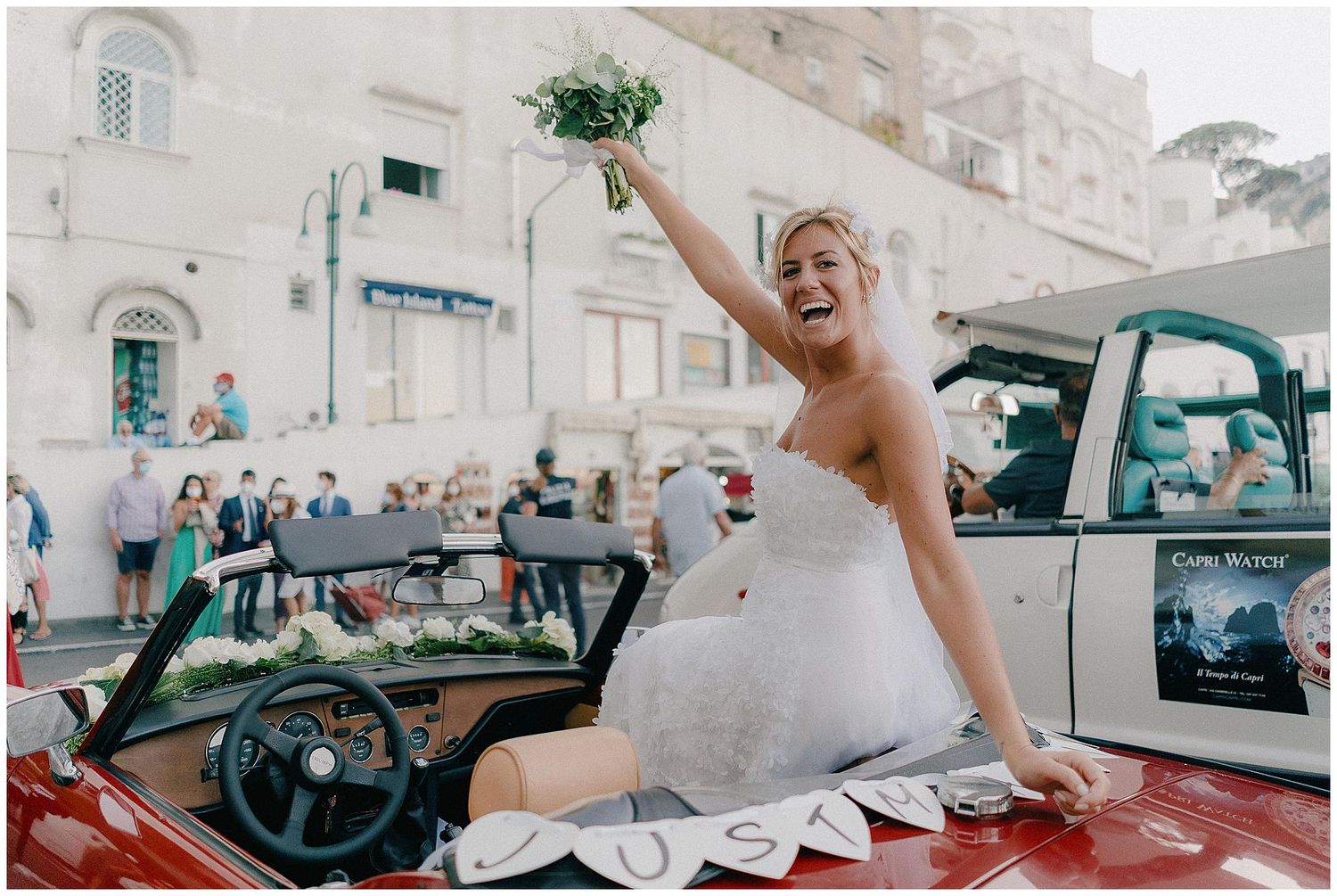 A joyful bride in a white wedding gown sits atop a vintage red car during a sunny celebration.