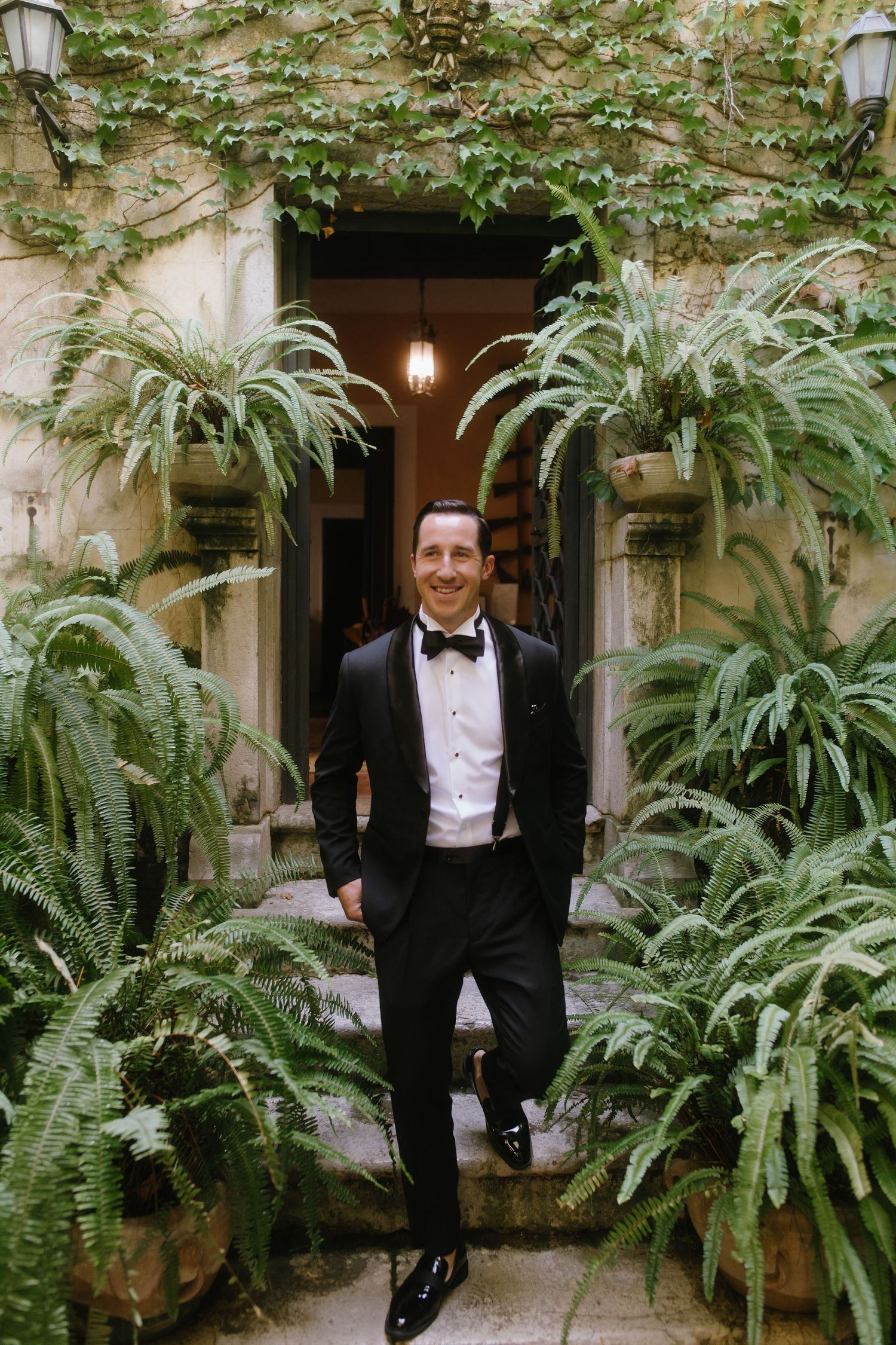 A person in a tuxedo poses in a lush garden courtyard with stone walls and hanging ferns.