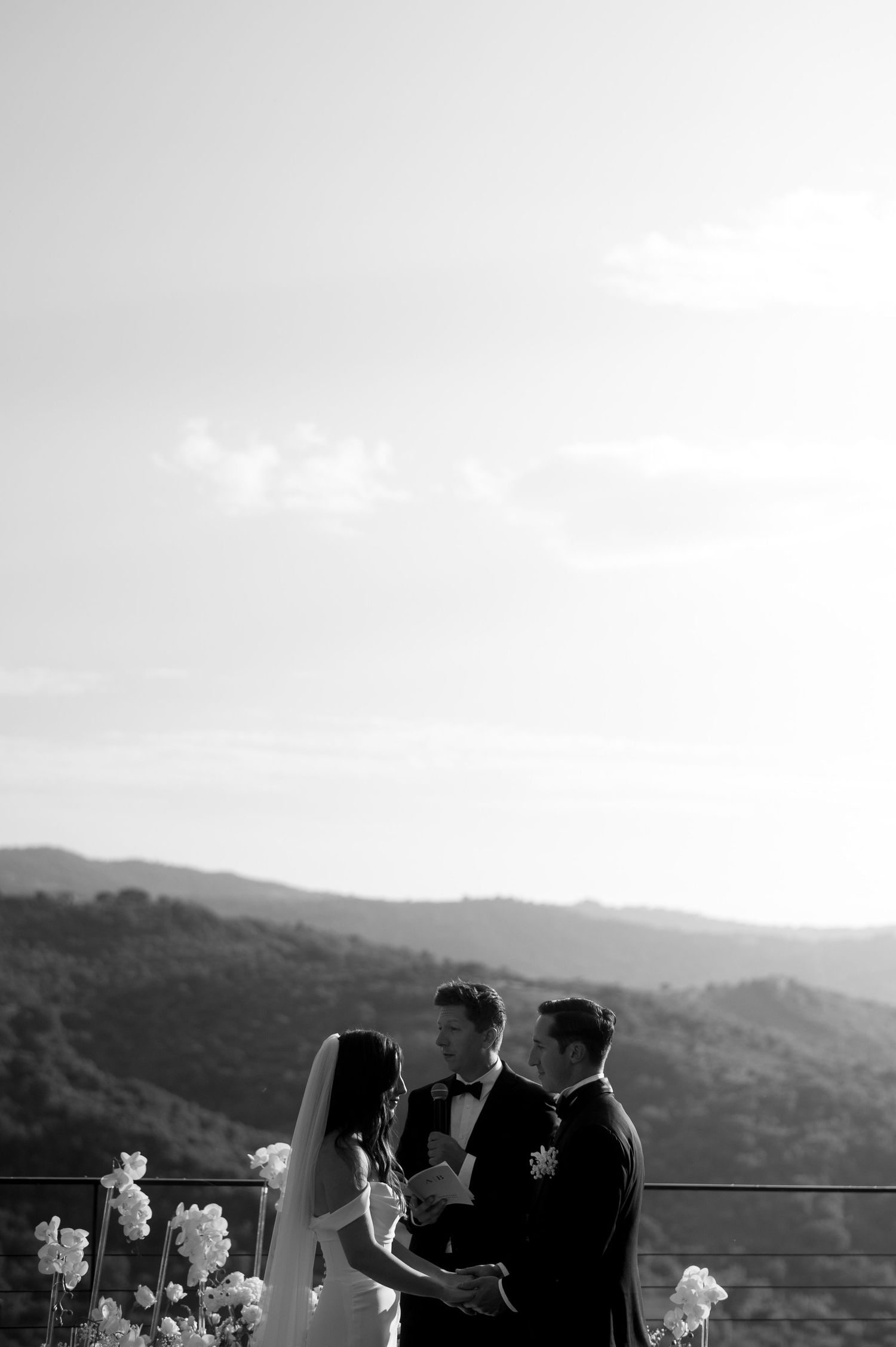 A scenic black and white wedding ceremony overlooking mountain ranges with guests gathered on an outdoor terrace.