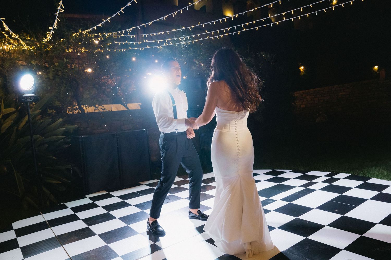 Couples dance together on a black and white checkered dance floor under twinkling string lights at an outdoor event.
